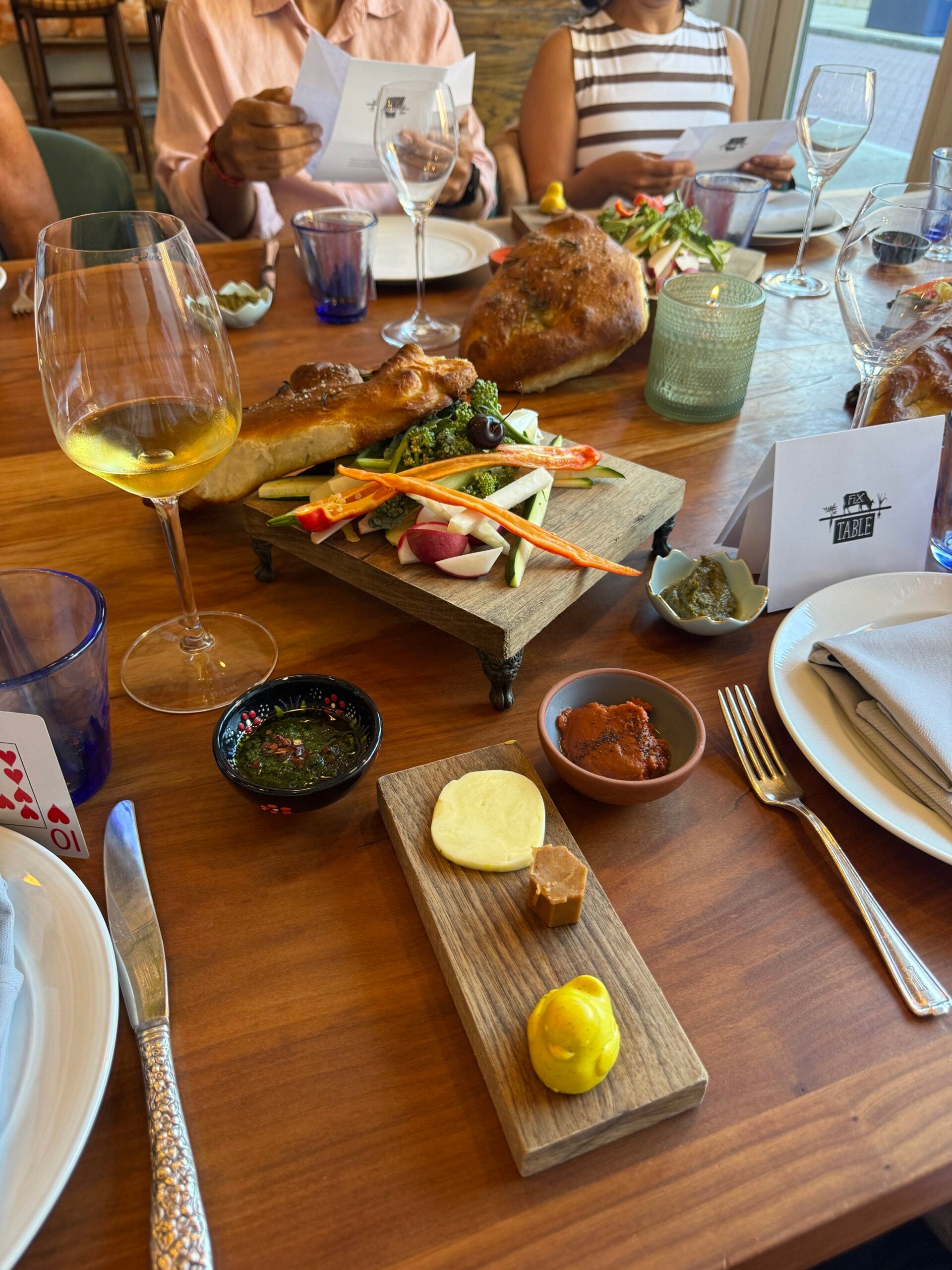 A wooden table set with wine, bread, veggies, dipping sauces, butter, and mustard. Three people are seated, with assorted glassware, plates, and menus, enjoying a meal together.