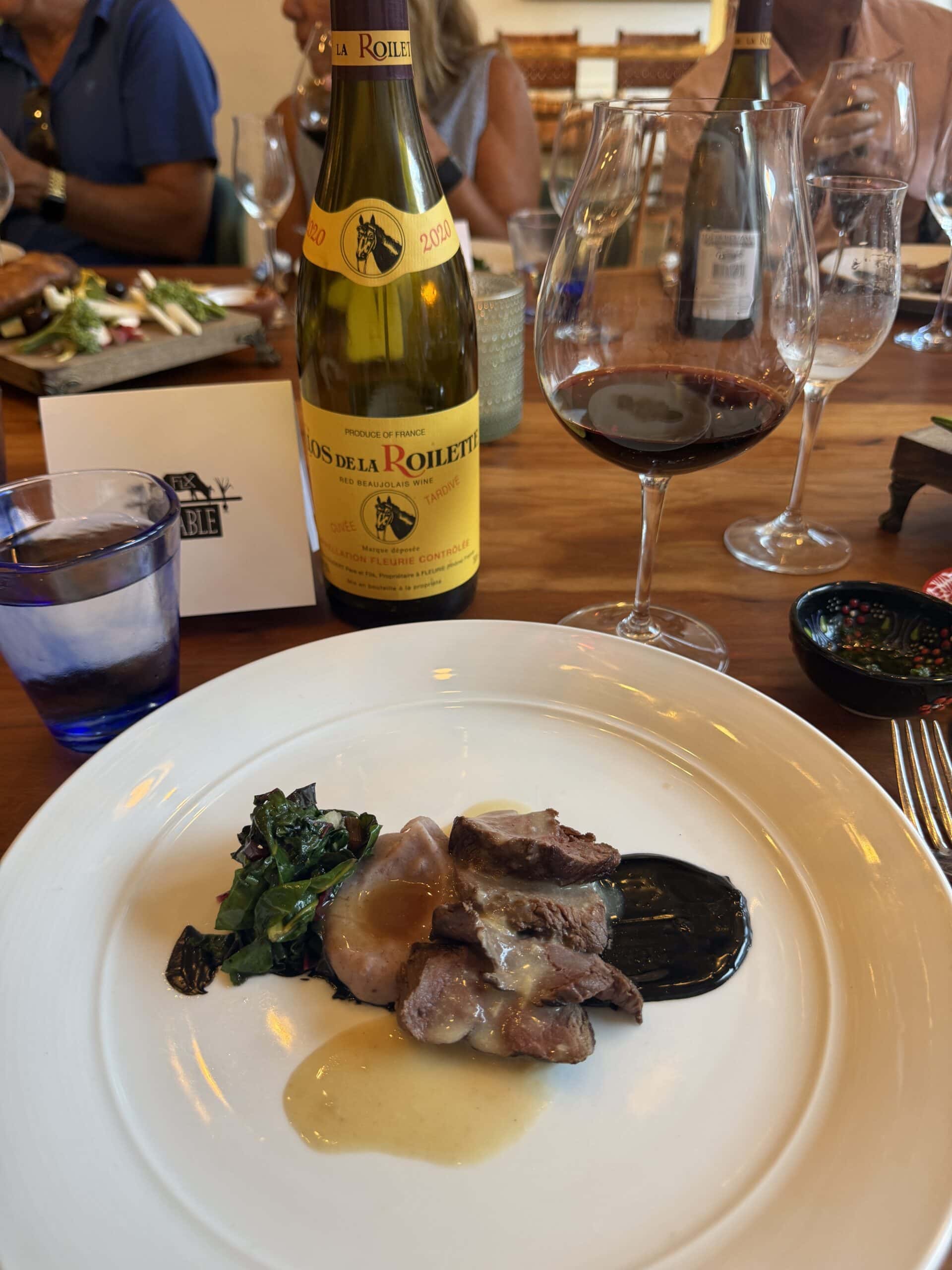 A plated dish with sliced meat, sauce, and greens sits on a table beside a wine glass, a bottle of red wine, and a water glass. Other diners and plates are visible in the background.