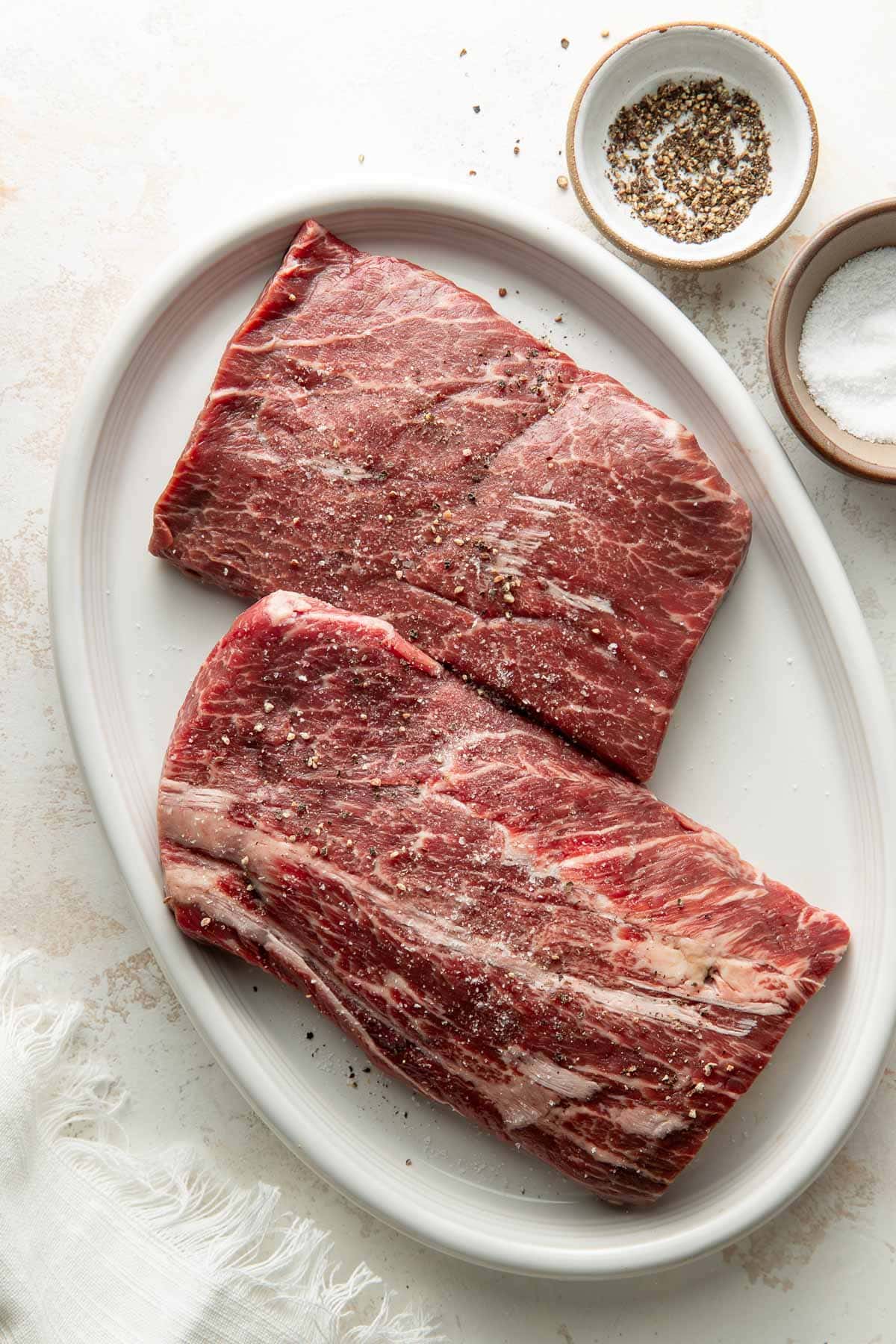 Two raw, seasoned pieces of steak on an oval white plate, with small bowls of salt and cracked black pepper nearby on a light surface.