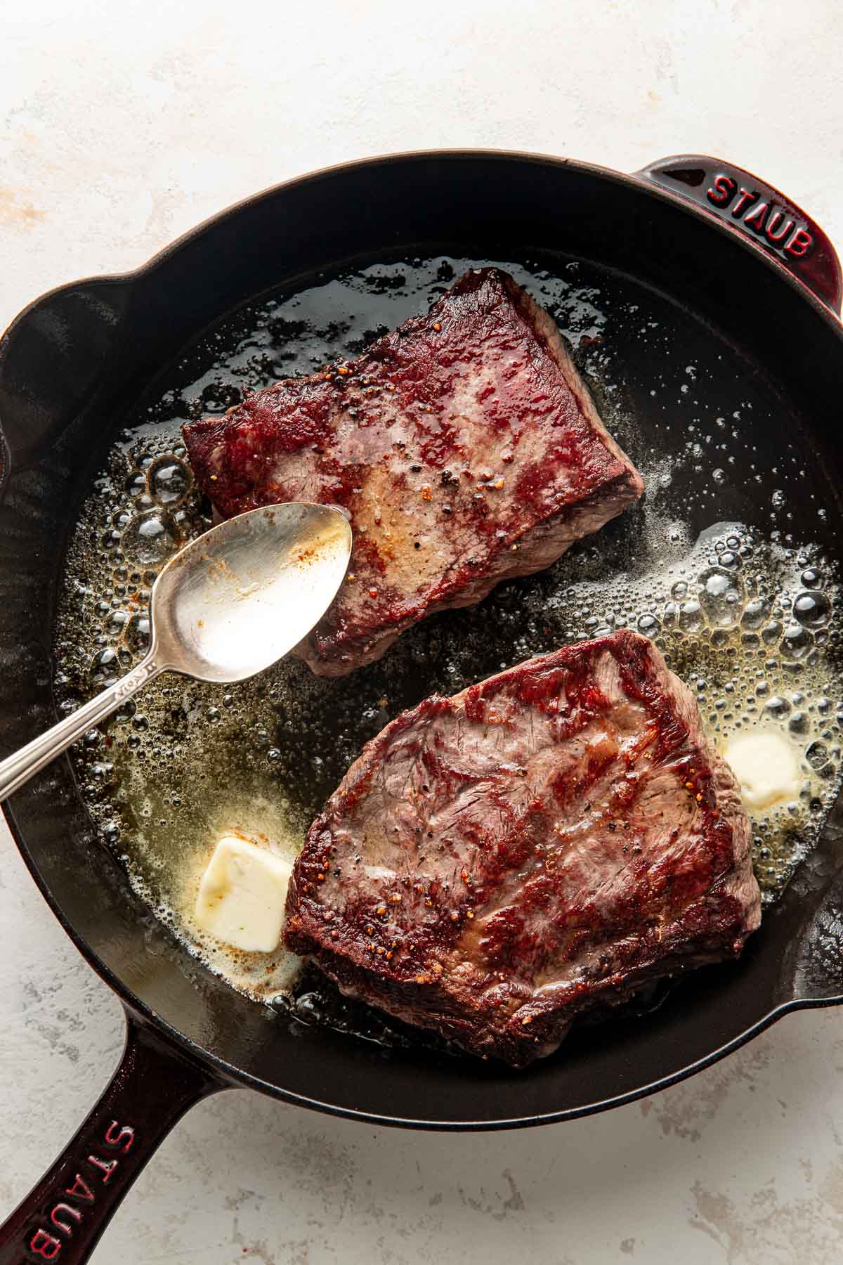 Two seared steaks in a cast iron skillet with bubbling butter and a spoon, viewed from above on a light surface.