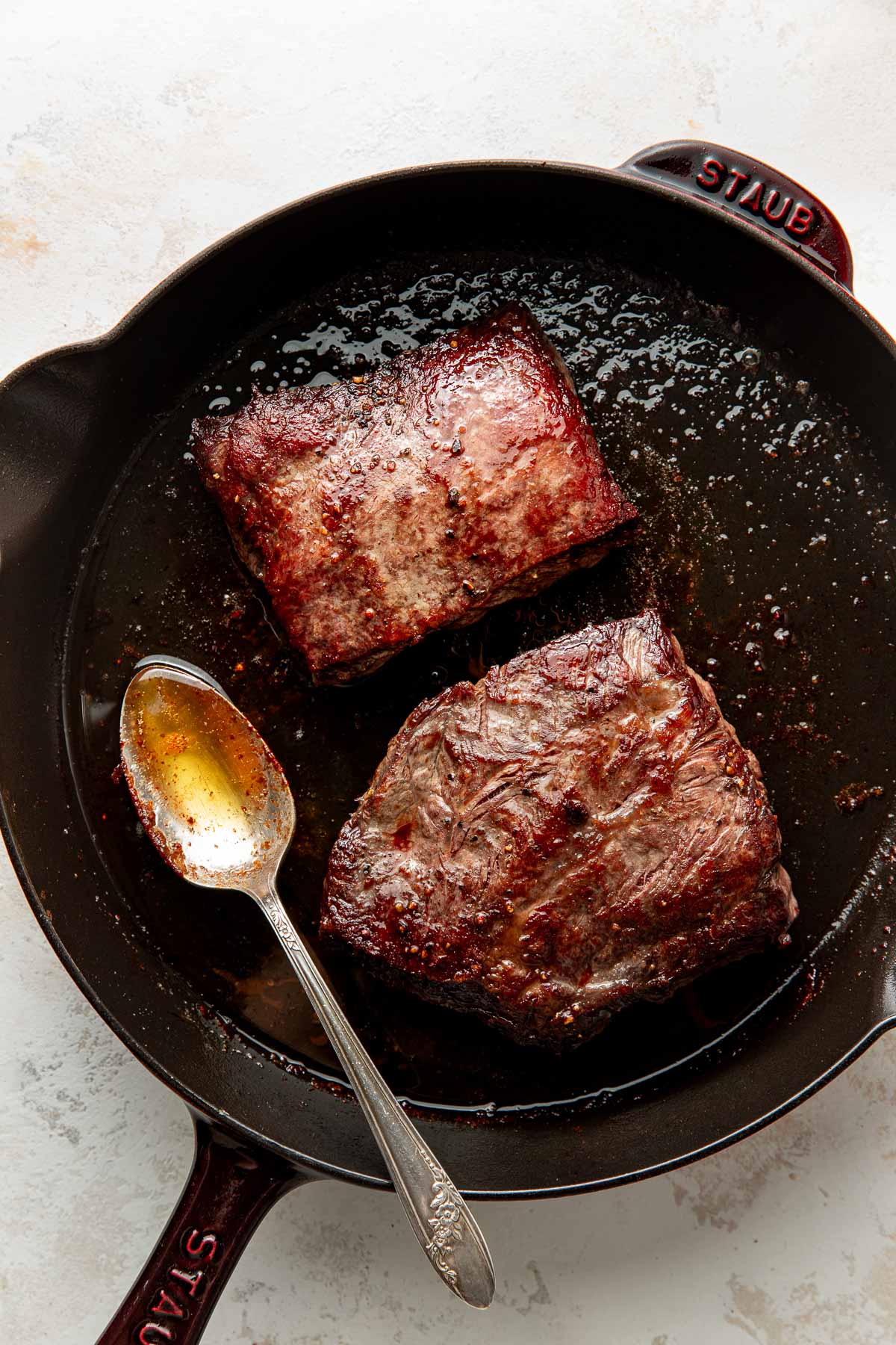 Two browned beef short ribs in a black cast iron skillet with a spoon resting beside them, containing some cooking juices.