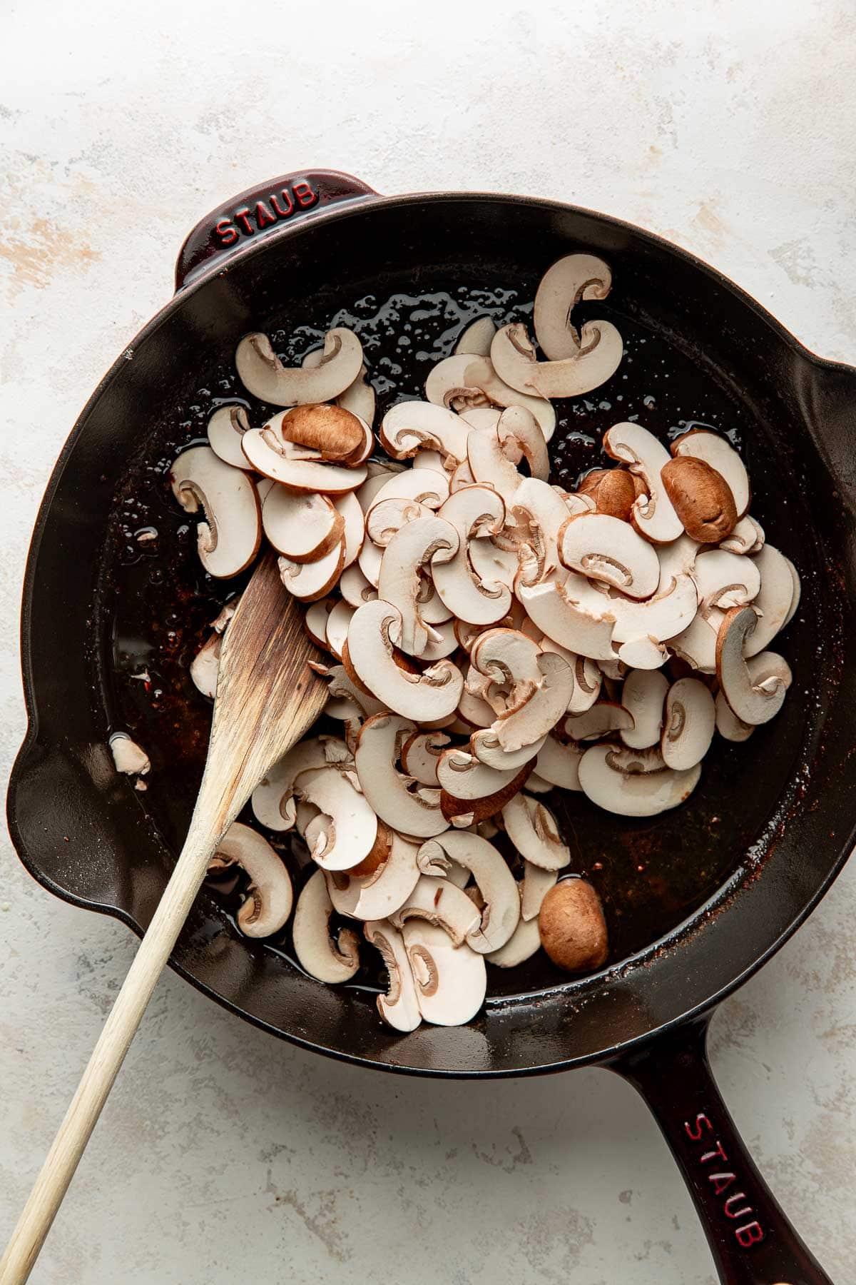 Sliced mushrooms in a black skillet with a wooden spoon, ready to be sautéed. Some mushrooms are whole, and the skillet sits on a light countertop.