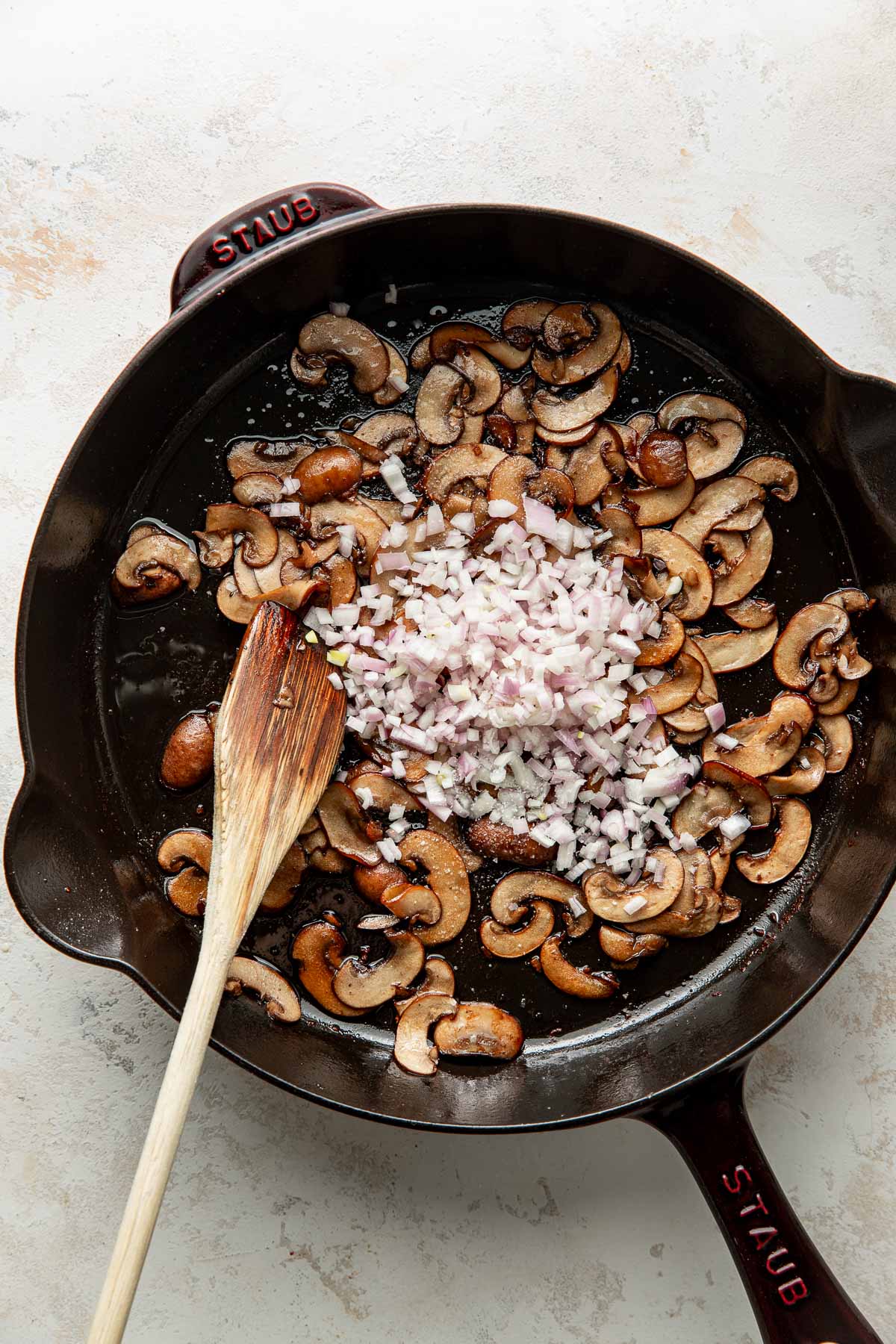 Sliced mushrooms and diced shallots cooking in a black skillet with a wooden spatula, on a light-colored countertop.