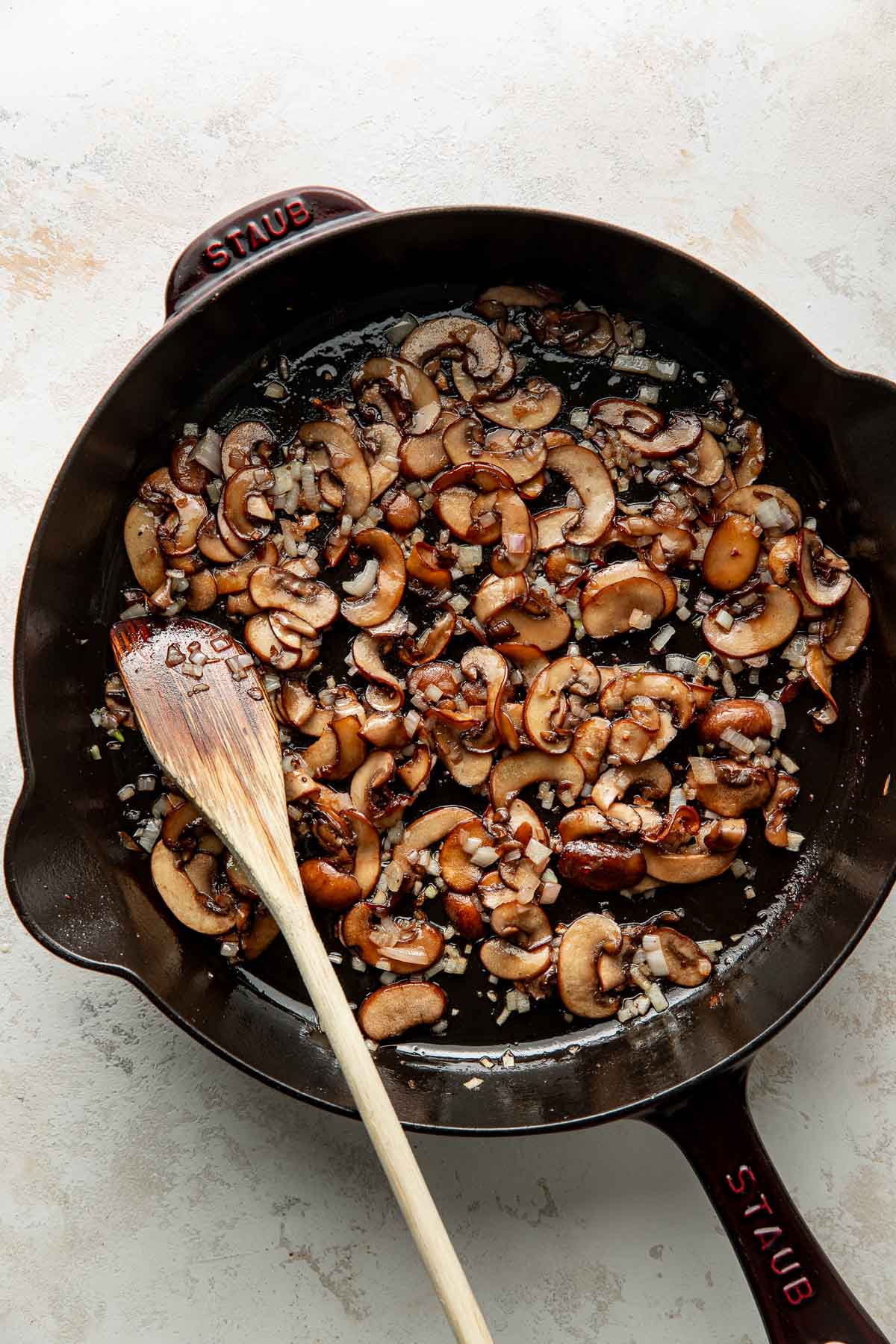 Sliced mushrooms and chopped onions sauté in a black cast iron skillet with a wooden spatula resting inside.