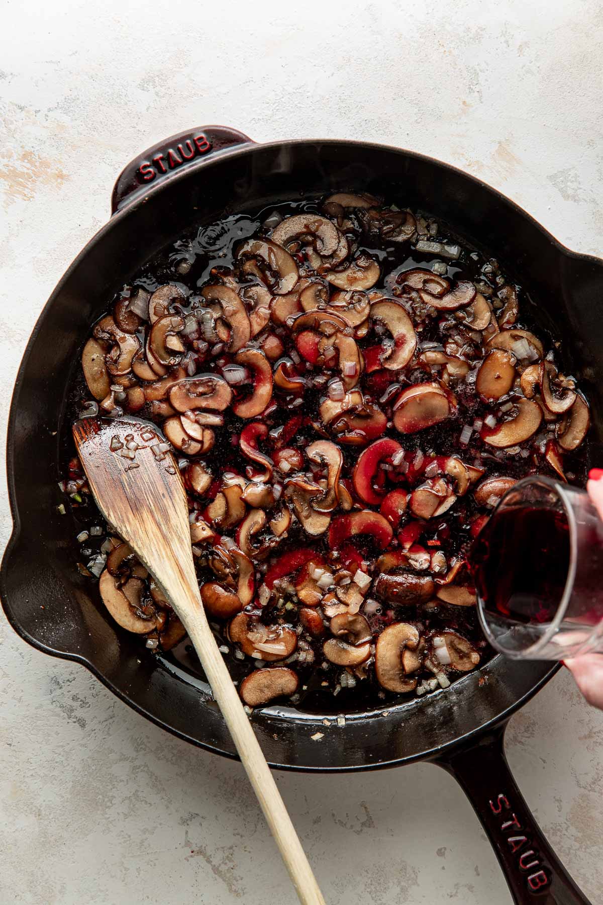 A cast iron skillet with sliced mushrooms, diced onions, and a wooden spoon, as red wine is being poured in.
