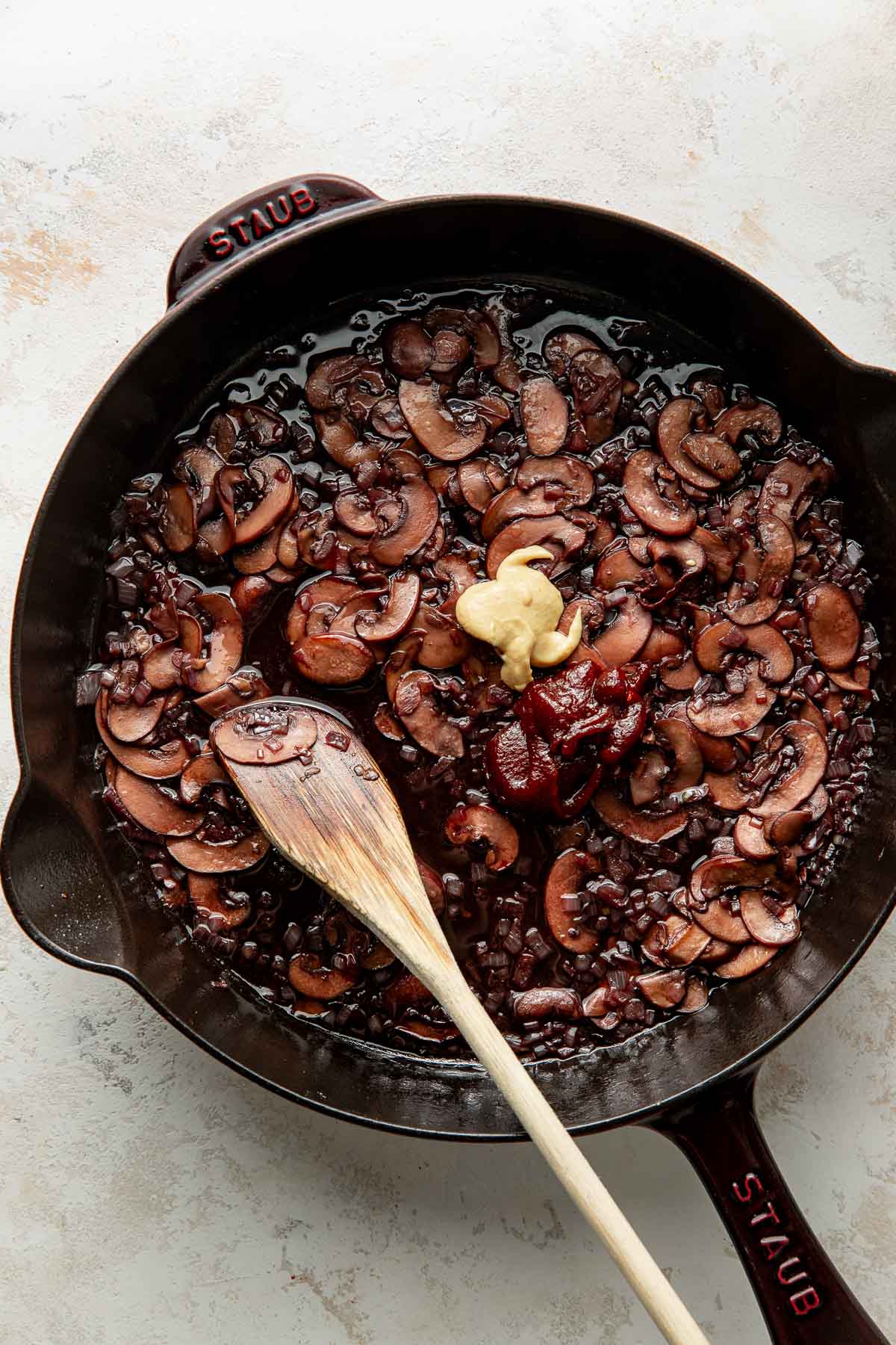 A cast iron skillet with sautéed sliced mushrooms, onions, tomato paste, and a dollop of creamy ingredient, with a wooden spoon resting inside.