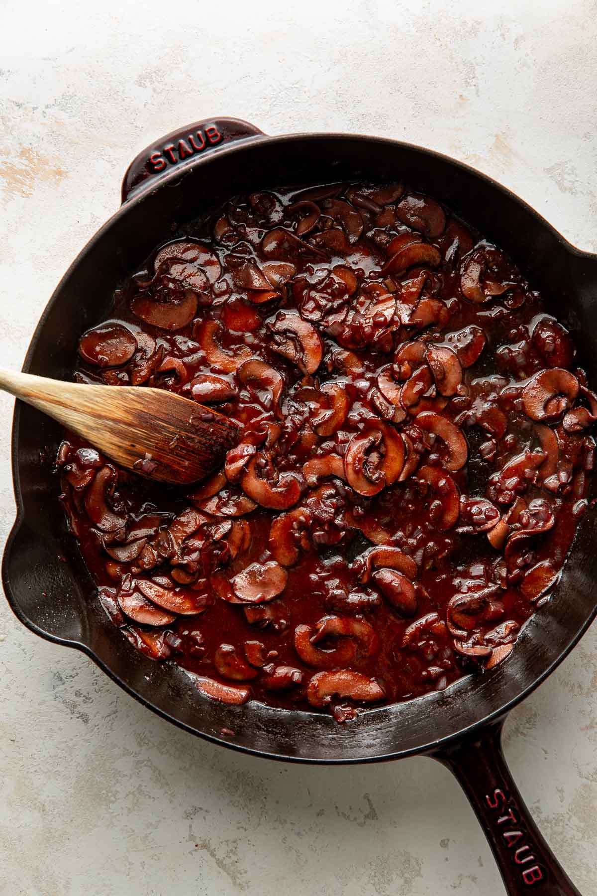 A cast iron skillet with sautéed mushrooms in a rich, dark sauce, stirred with a wooden spoon on a light textured surface.