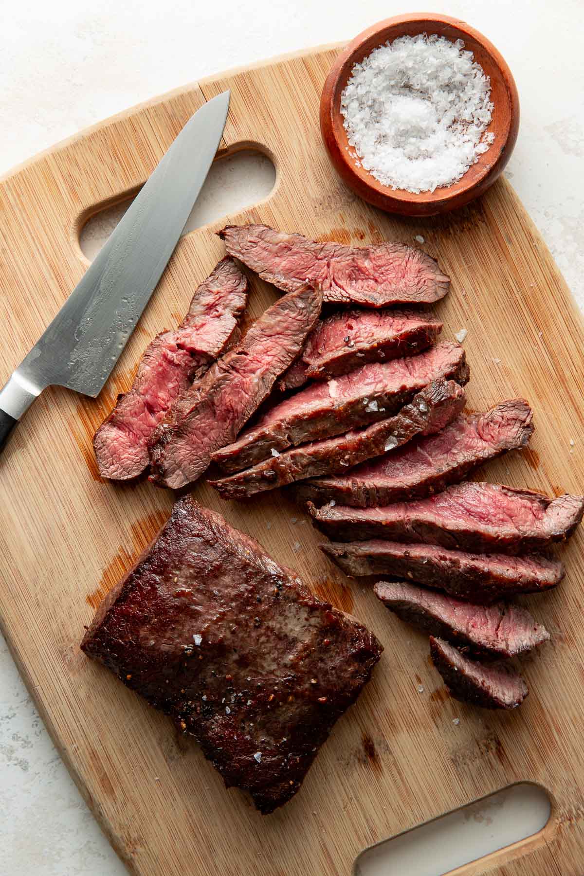 Sliced medium-rare steak on a wooden cutting board with a knife and a small bowl of coarse salt nearby.