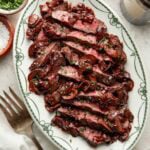 Sliced steak topped with mushrooms and sauce on an oval platter, garnished with chopped chives. A bowl of salt, chopped herbs, a fork, and a glass of red wine are nearby.