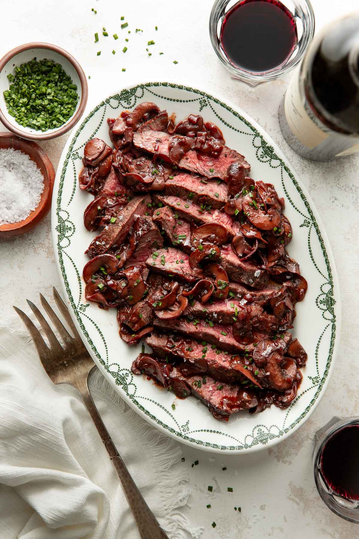 Sliced steak with mushrooms and herbs is arranged on a decorative oval plate, surrounded by small bowls of salt and chopped chives, a white napkin, a large fork, and two glasses of red wine.