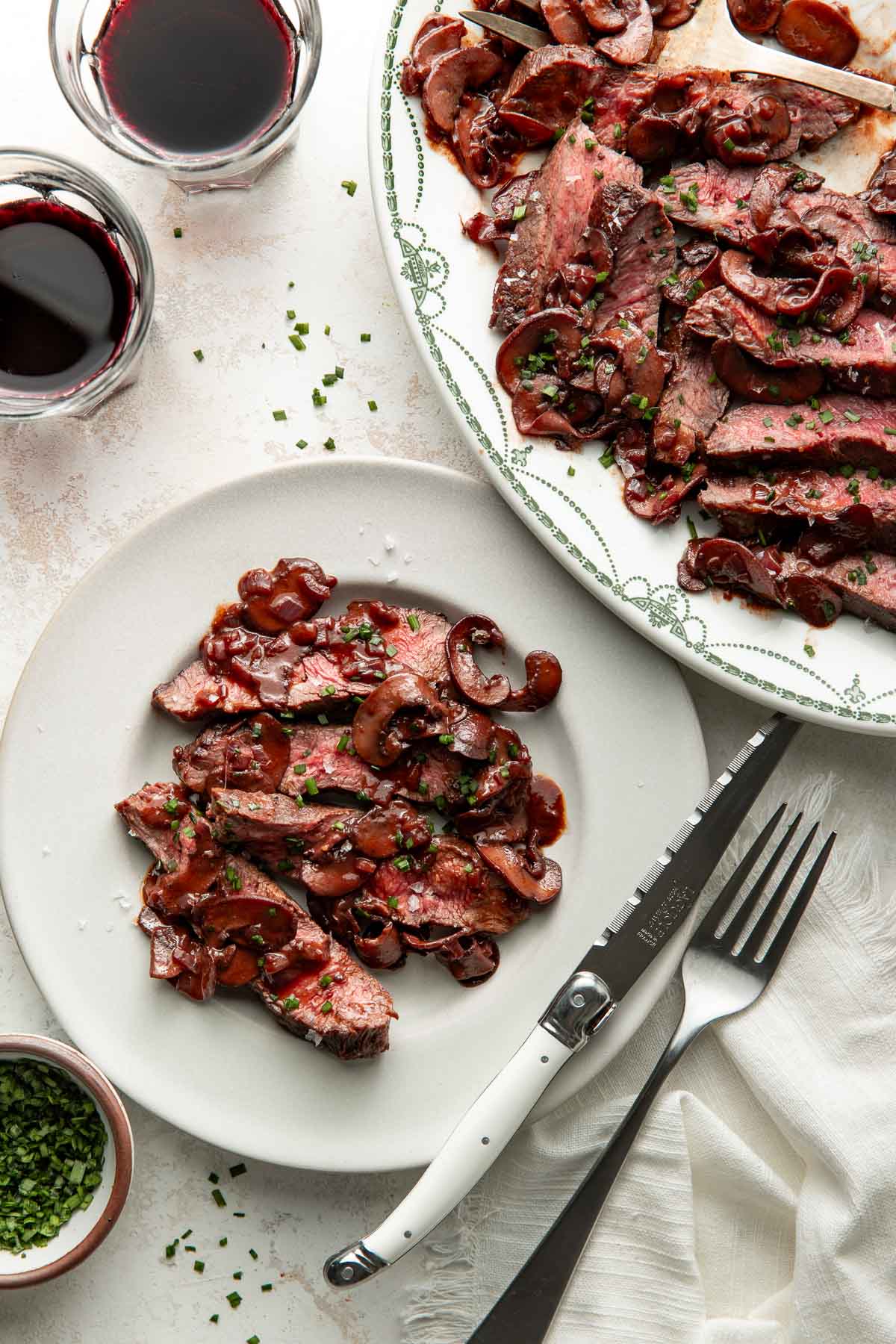 Sliced steak topped with mushroom sauce on a white plate, garnished with herbs, with a fork and knife beside it. A larger platter of steak and glasses of red wine are also visible.