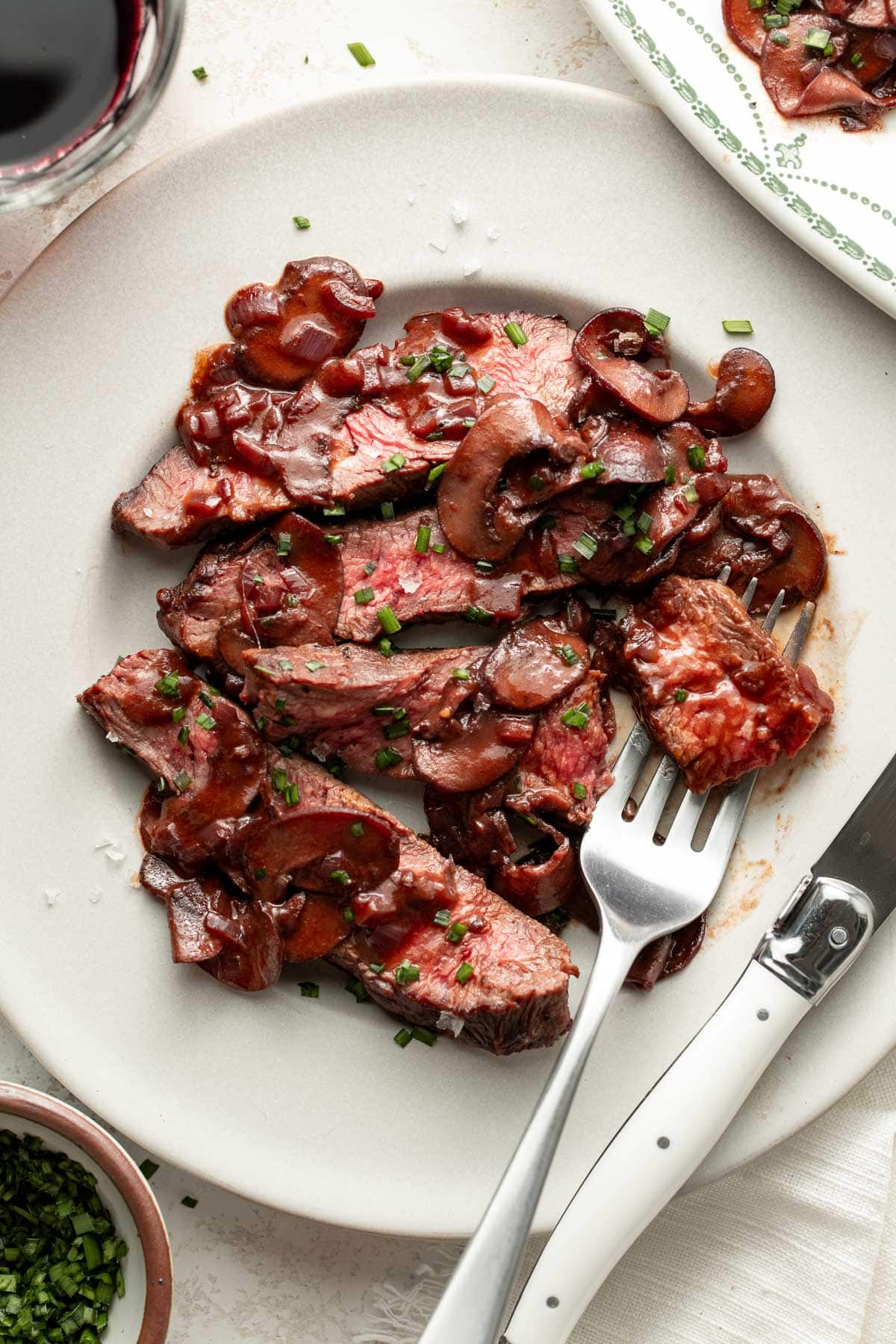 Sliced steak topped with a mushroom and red wine sauce, garnished with chopped chives, is served on a white plate with a fork and knife.