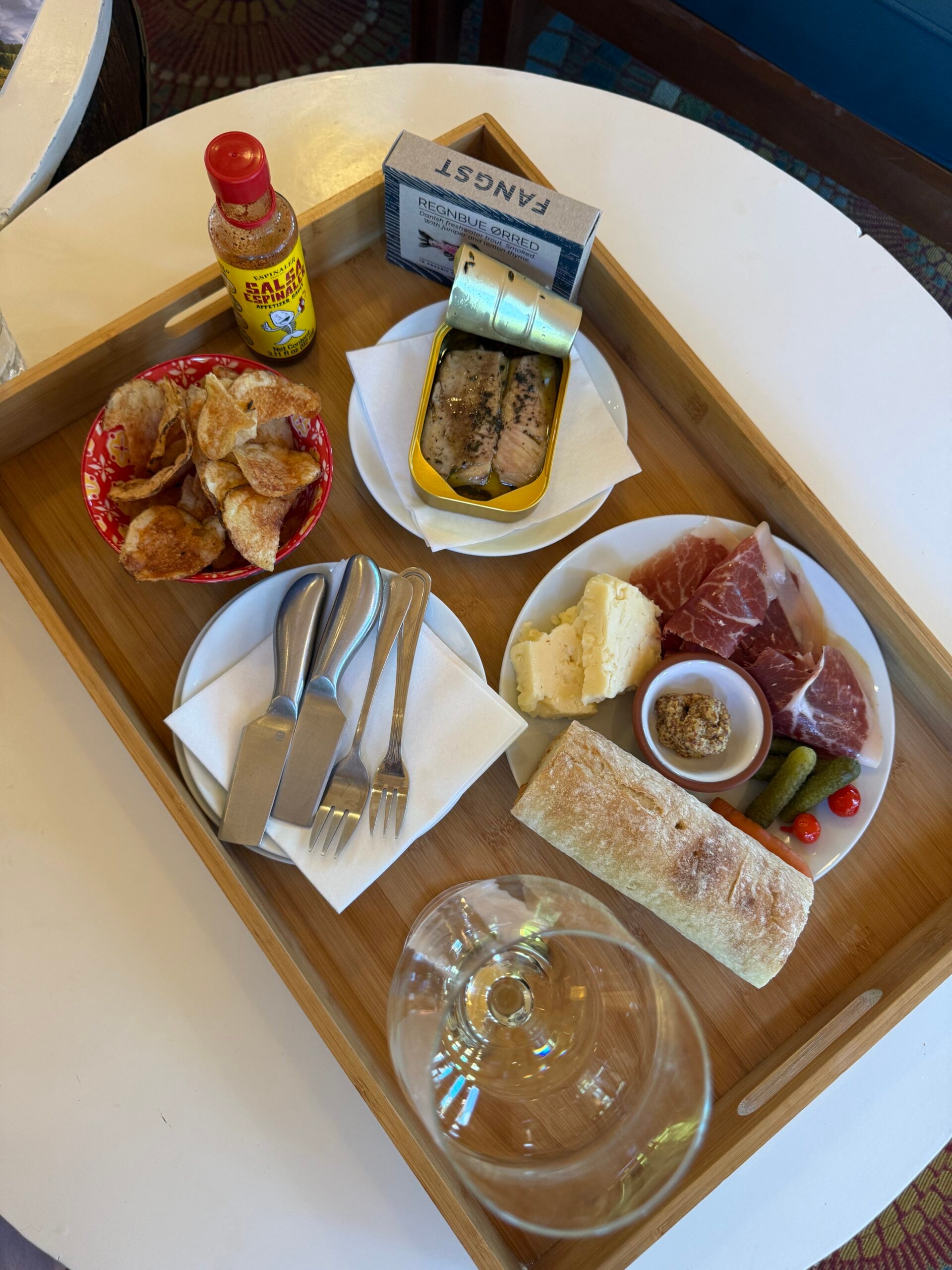A wooden tray with chips, canned fish, cheese, prosciutto, bread, pickles, cherry tomatoes, mustard, a bottle of hot sauce, forks, napkins, and an empty wine glass.
