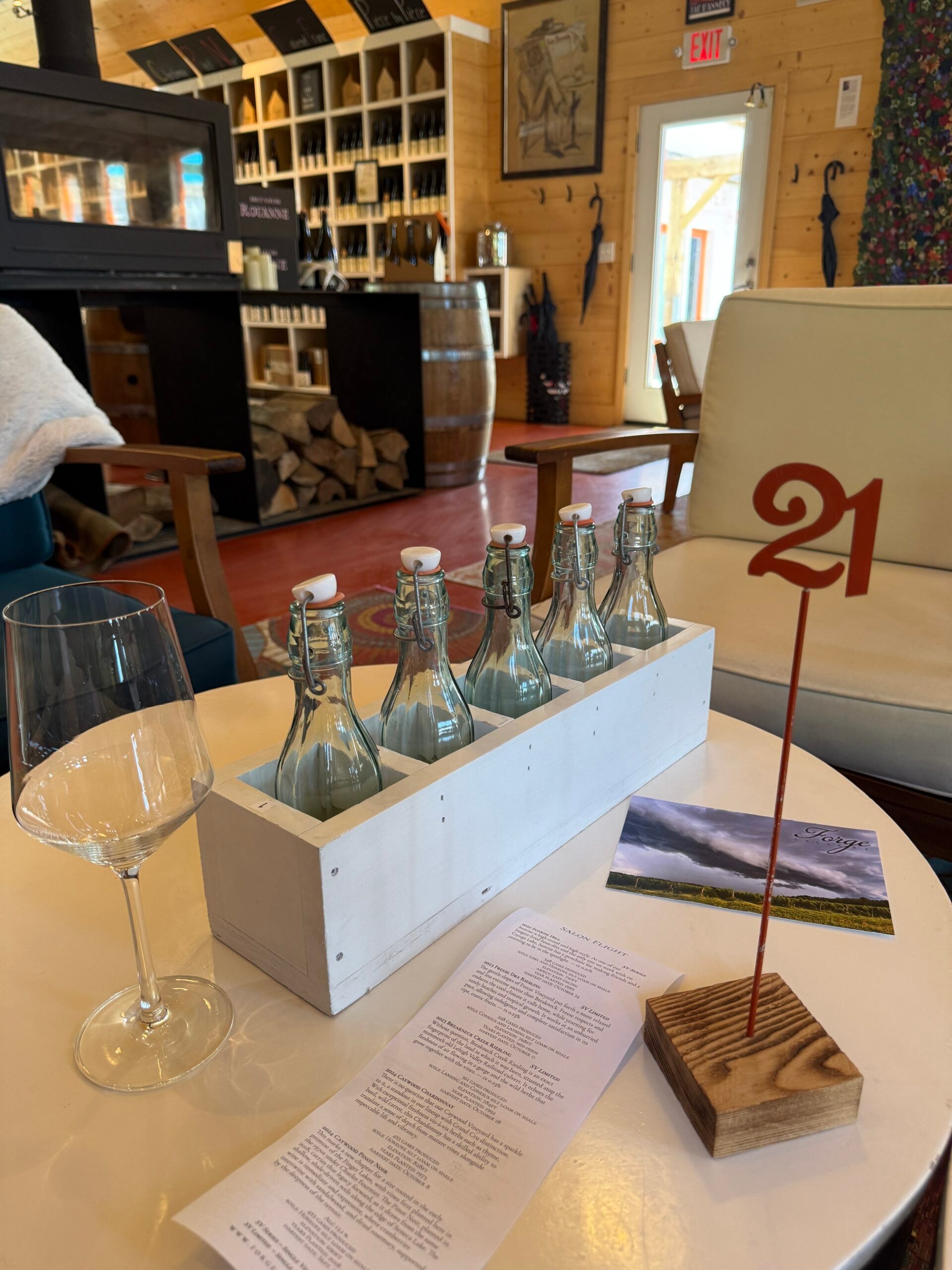 A round table with a glass, a menu, five bottled drinks in a white holder, and a table number 21. The cozy room has wooden walls, wine bottles, a barrel, firewood, and soft seating in the background.