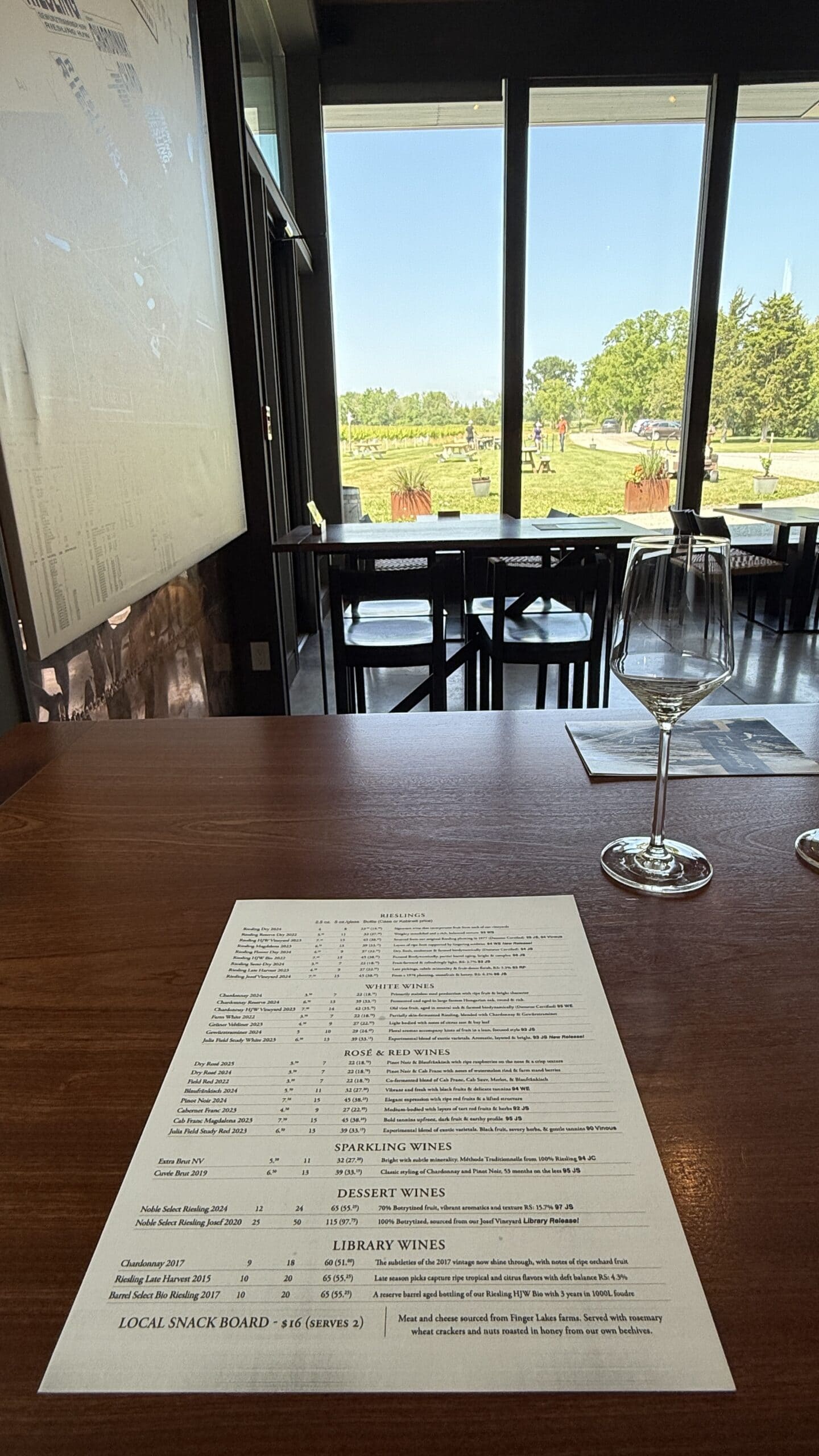 A wine tasting menu sits on a wooden table next to an empty wine glass. Large windows reveal a sunny outdoor scene with green grass, trees, and people in the distance.