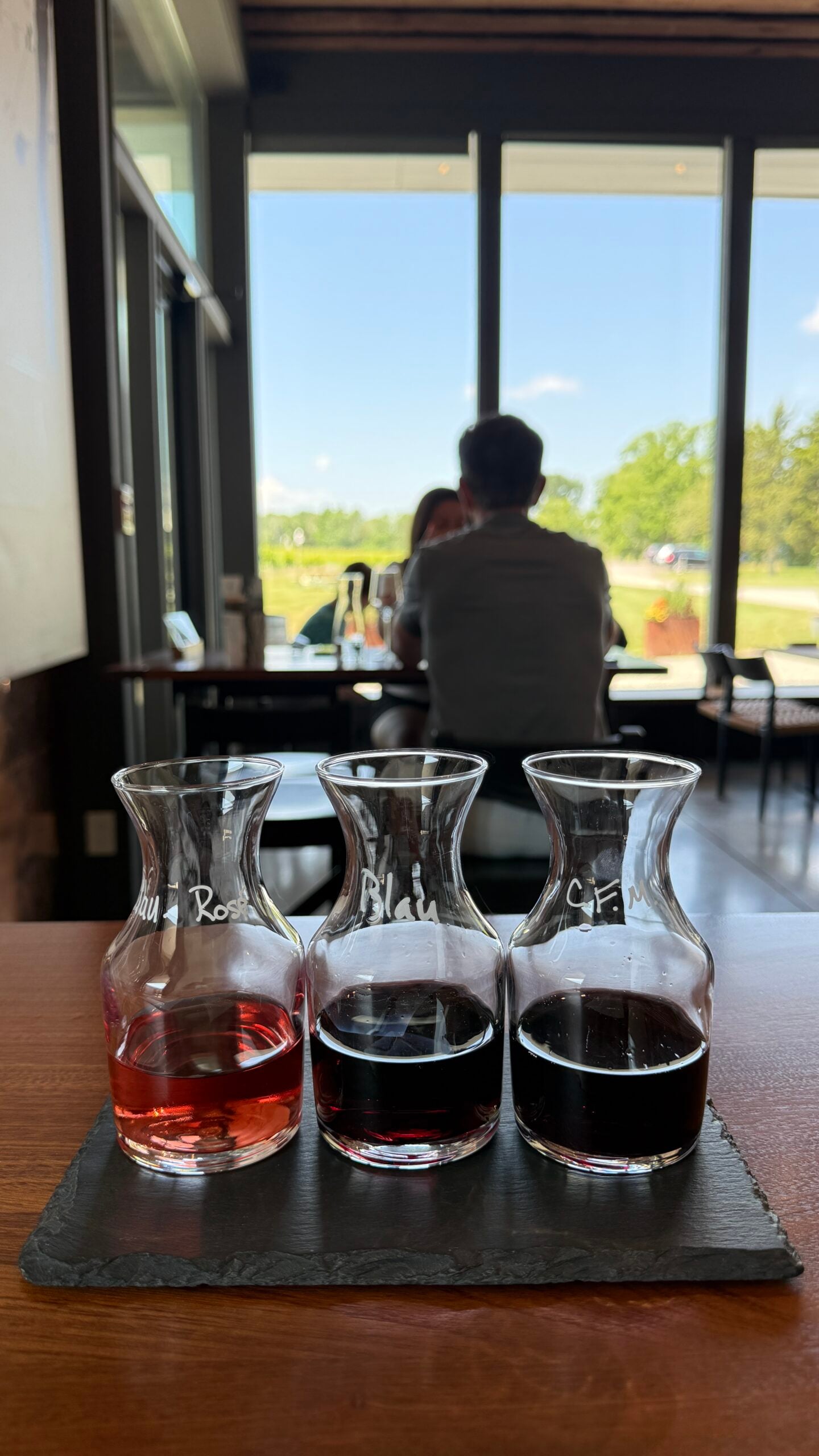Three small glass carafes with red wines on a slate, set on a wooden table. A couple sits by large windows with a sunny view of trees and grass in the background.