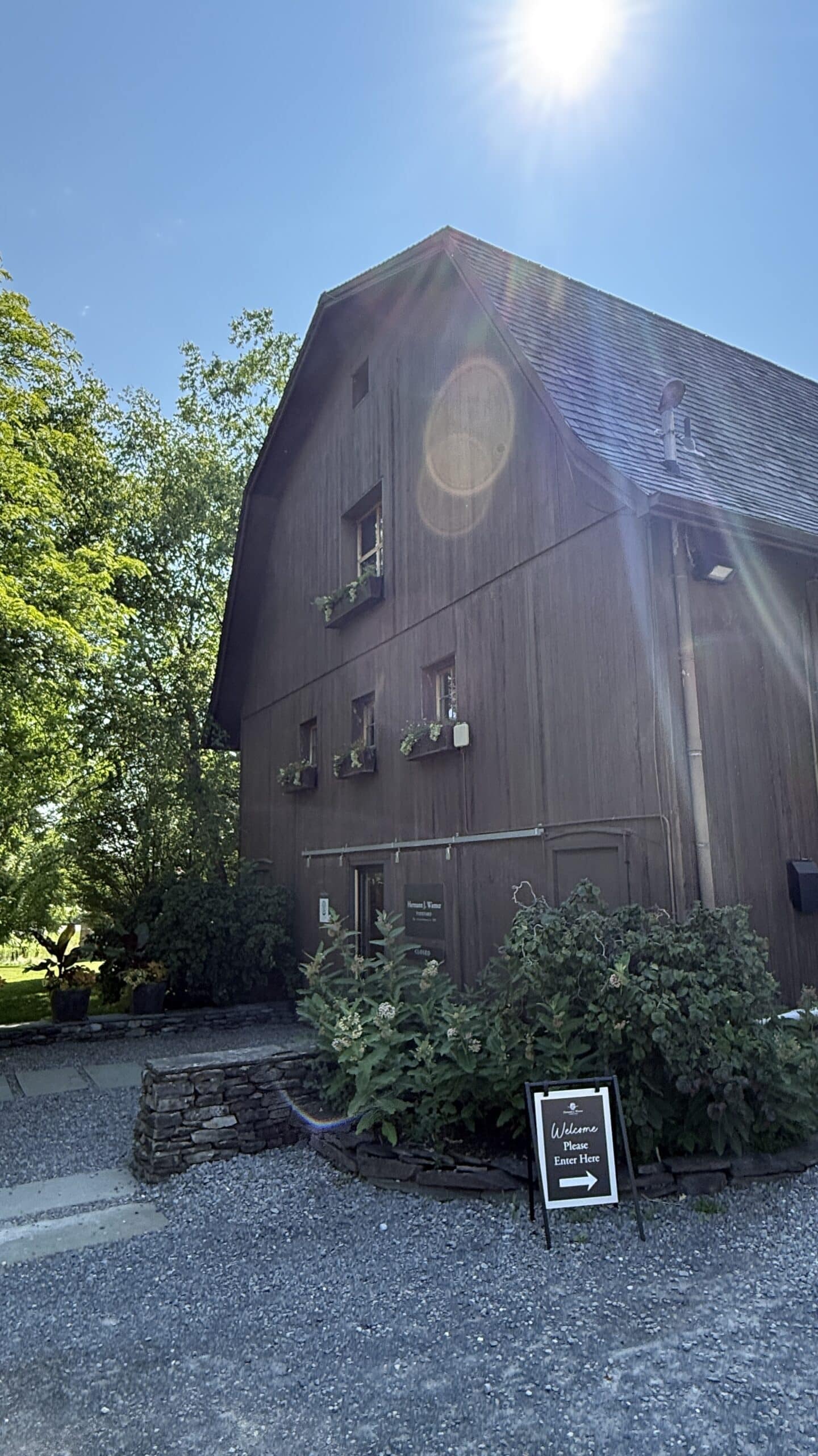 A rustic wooden building with flower boxes on the windows sits under a sunny sky. A sign near the entrance welcomes visitors, and trees provide shade along the gravel path.