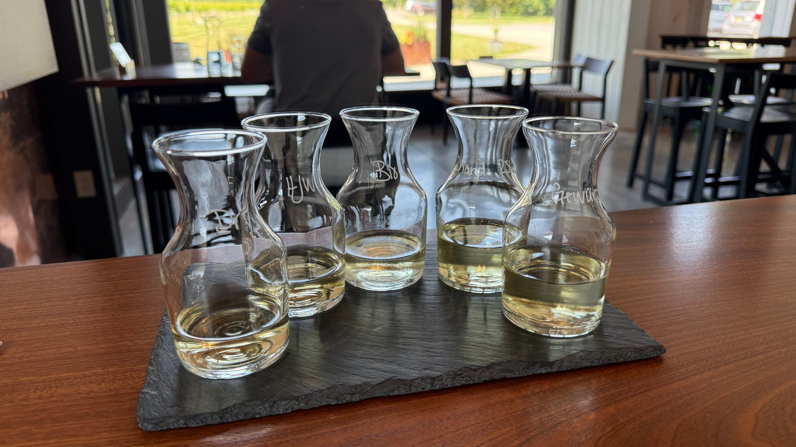 Five small glass carafes with light-colored liquid are arranged on a black slate tray on a wooden table in a bright, modern cafe setting.