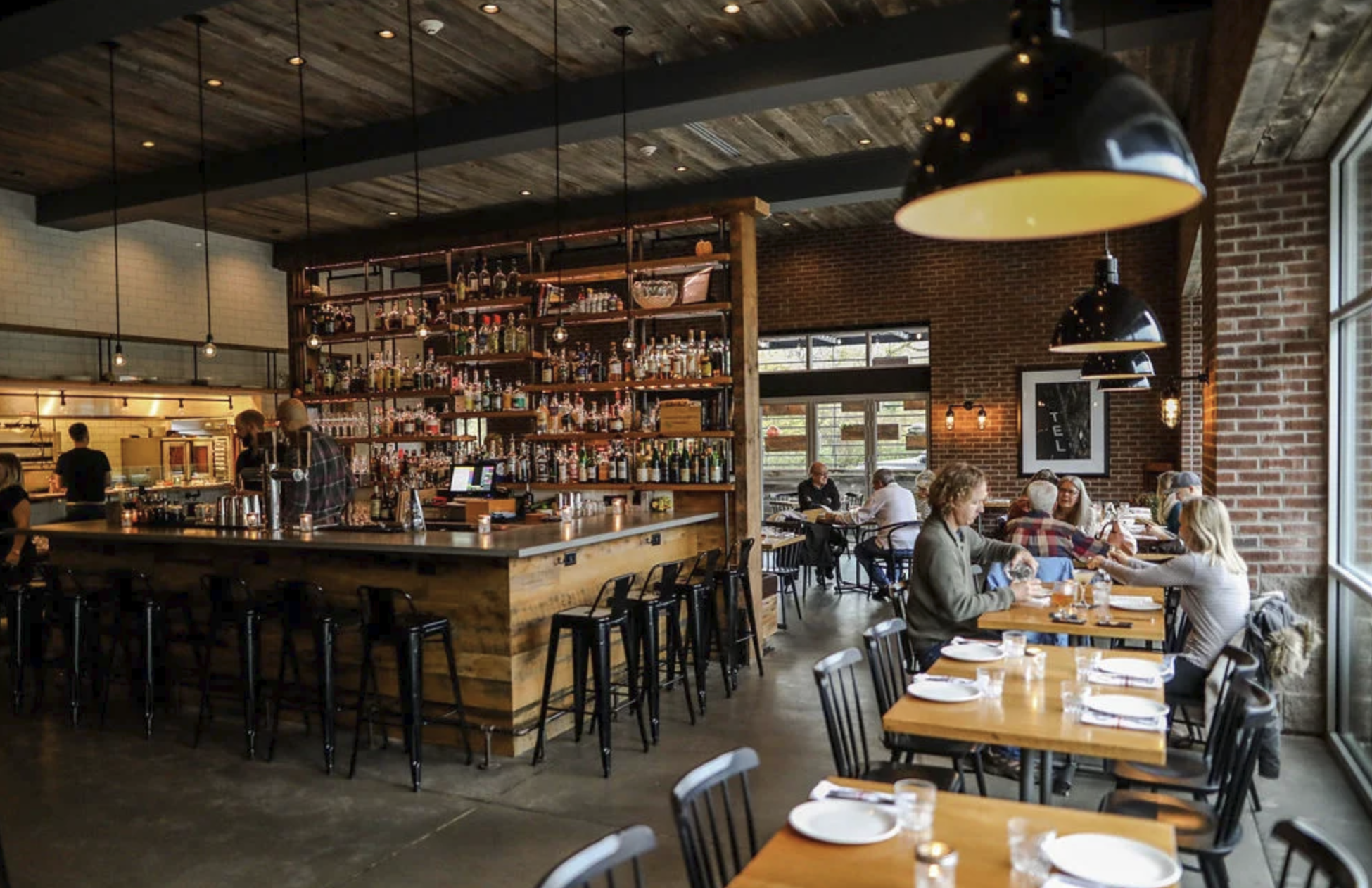 Modern restaurant interior with a wooden bar, high shelves of liquor, and pendant lights reflects the warm hospitality found in the Finger Lakes region. Exposed brick walls and large windows create an inviting atmosphere where people dine at tables, evoking the relaxed charm of a Finger Lakes getaway.