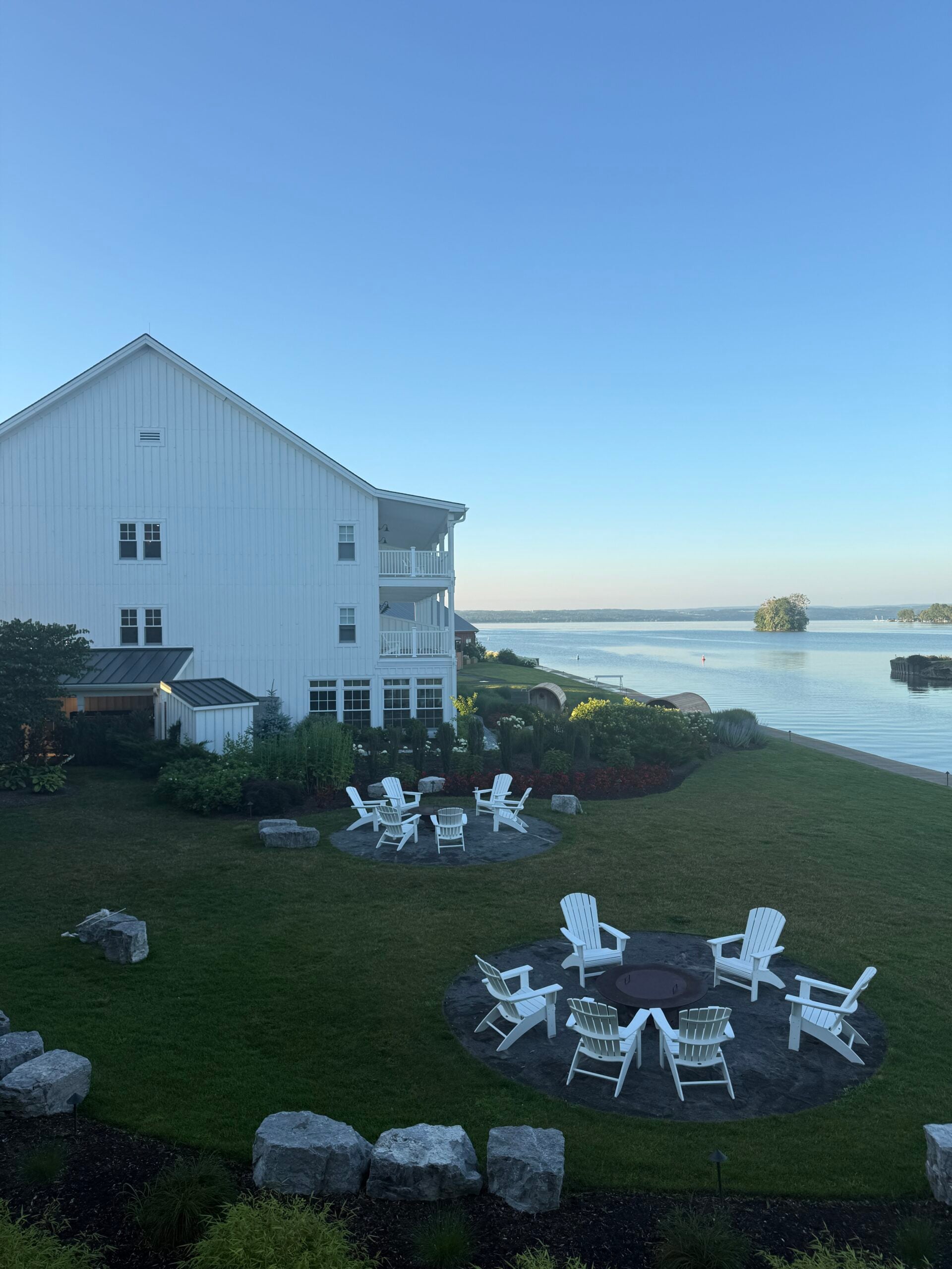 White lakeside building with balconies overlooks a green lawn with white Adirondack chairs arranged around two fire pits. Calm water and small islands are visible in the background under a clear blue sky.