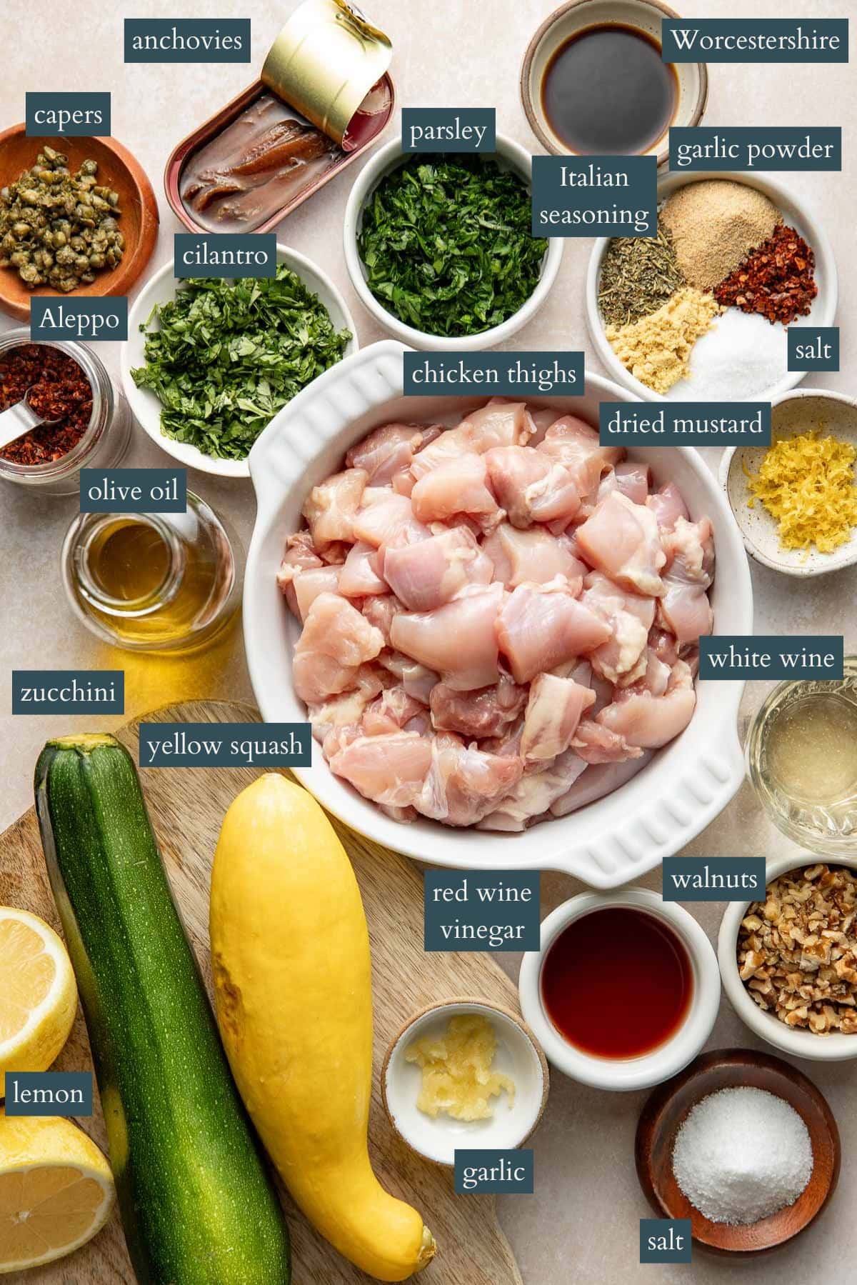A top-down view of various labeled ingredients in bowls and on a surface, including chopped chicken thighs, herbs, capers, anchovies, spices, walnuts, zucchini, yellow squash, lemon, olive oil, and vinegar.