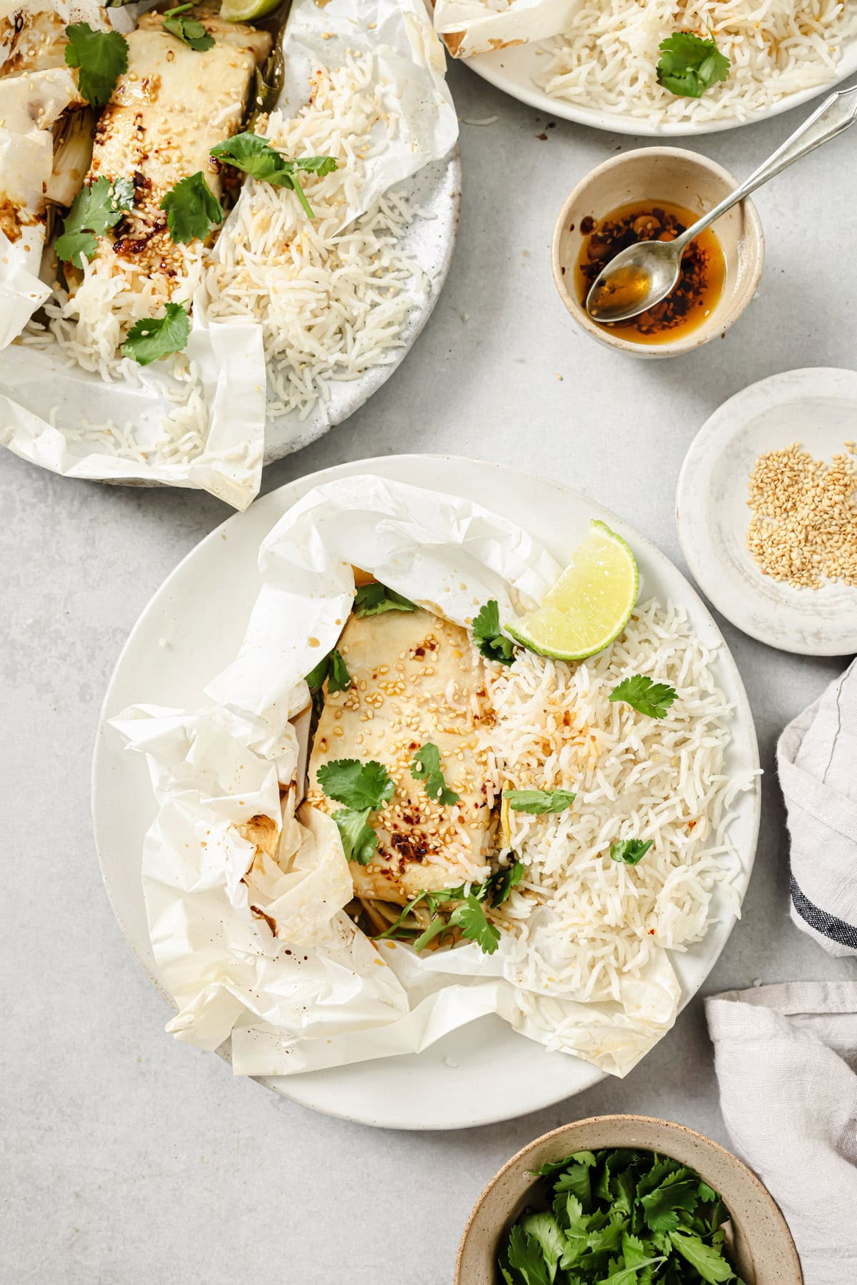 A plate with baked fish in parchment paper, served with white rice, garnished with cilantro and a lime wedge. Nearby are a bowl of sesame seeds, a small bowl of sauce with a spoon, and a bowl of fresh cilantro.