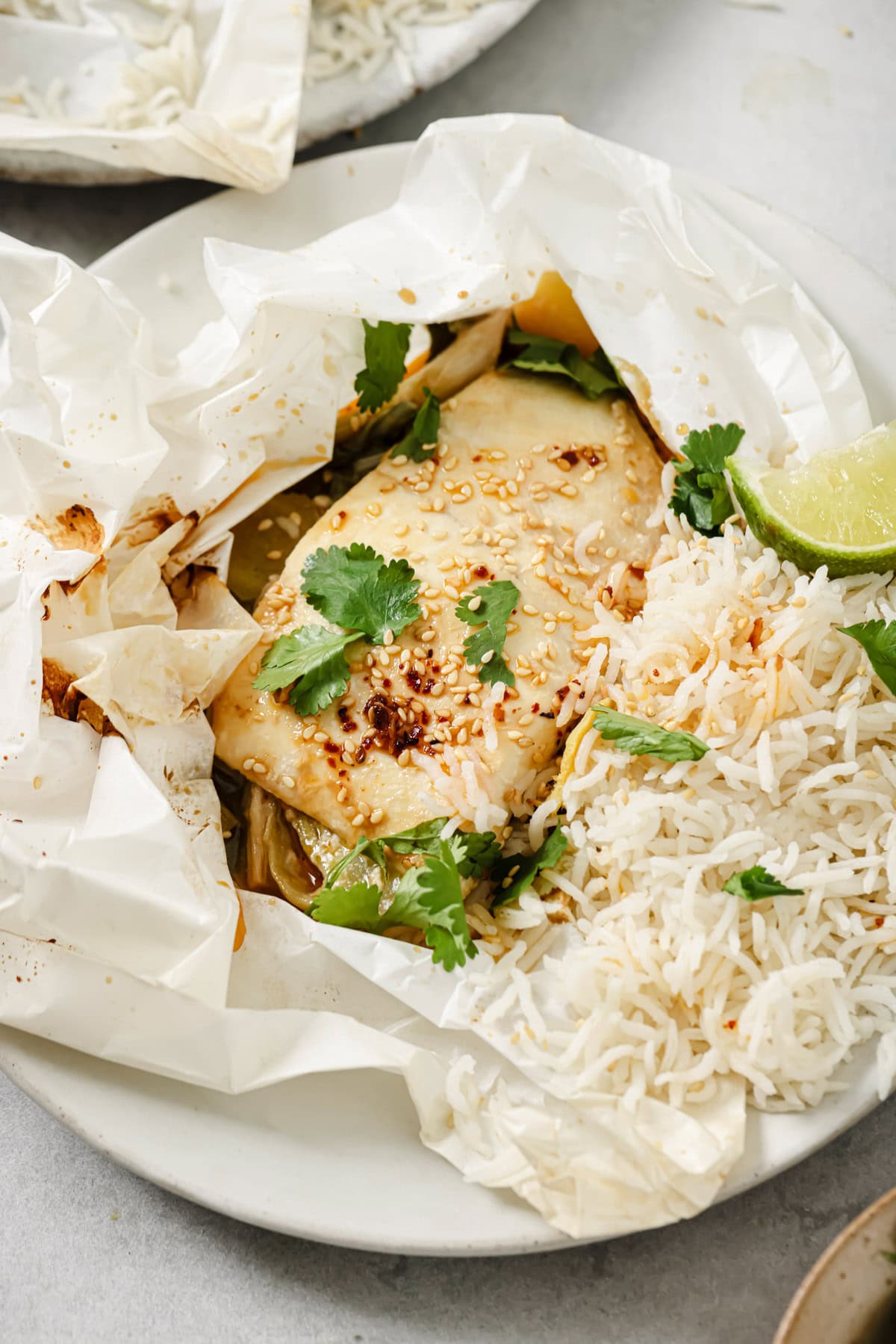 A close-up of fish baked in parchment paper, topped with sesame seeds and fresh cilantro, served with white rice and a lime wedge on a white plate.