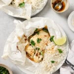 An overhead view of fish baked in parchment paper served with white rice, garnished with cilantro and a lime wedge. Small bowls of sauce, herbs, and a spoon are nearby on a light surface.