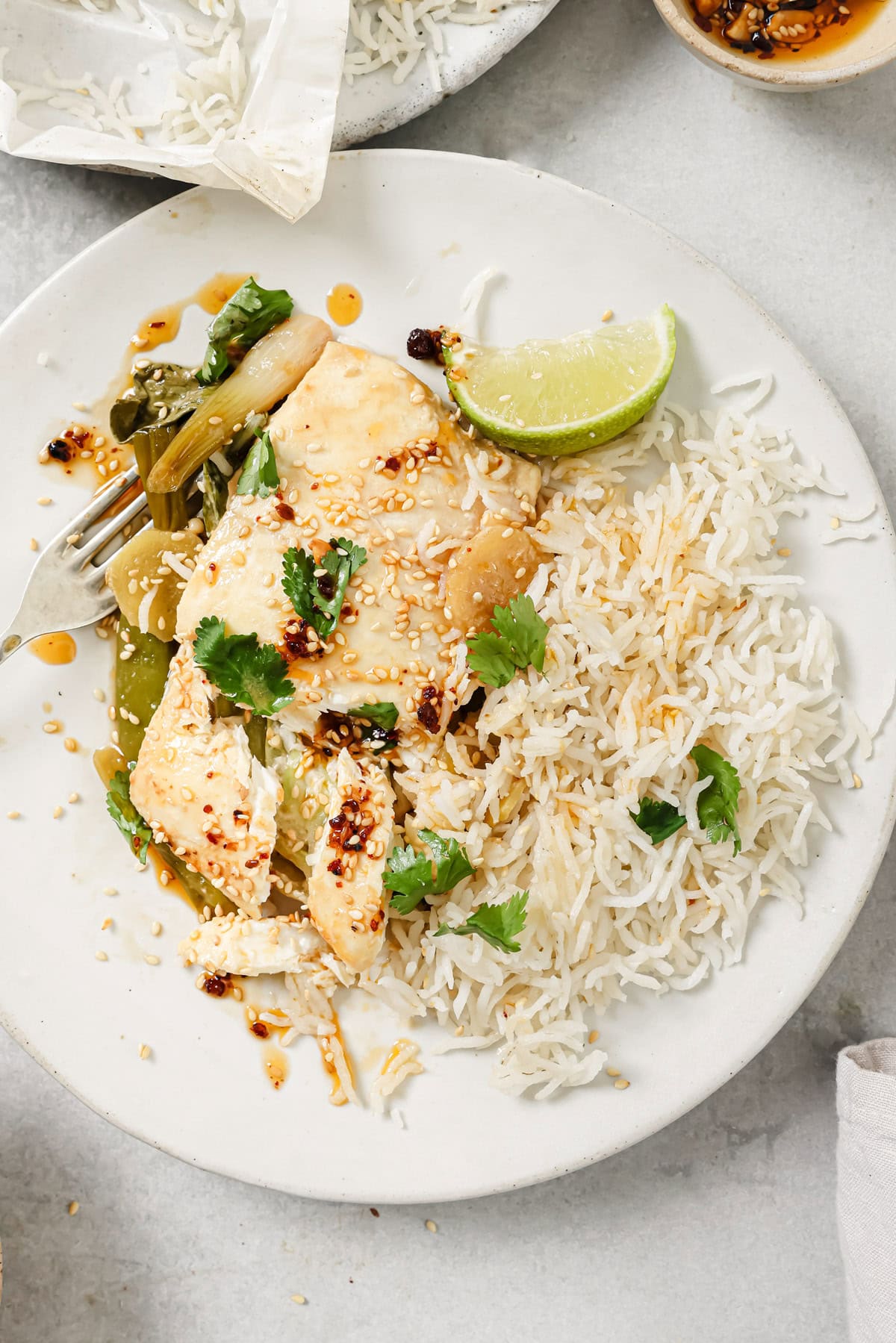A plate with white rice, steamed fish topped with cilantro, sesame seeds, and chili oil, sautéed vegetables, and a lime wedge, with a fork on the side.