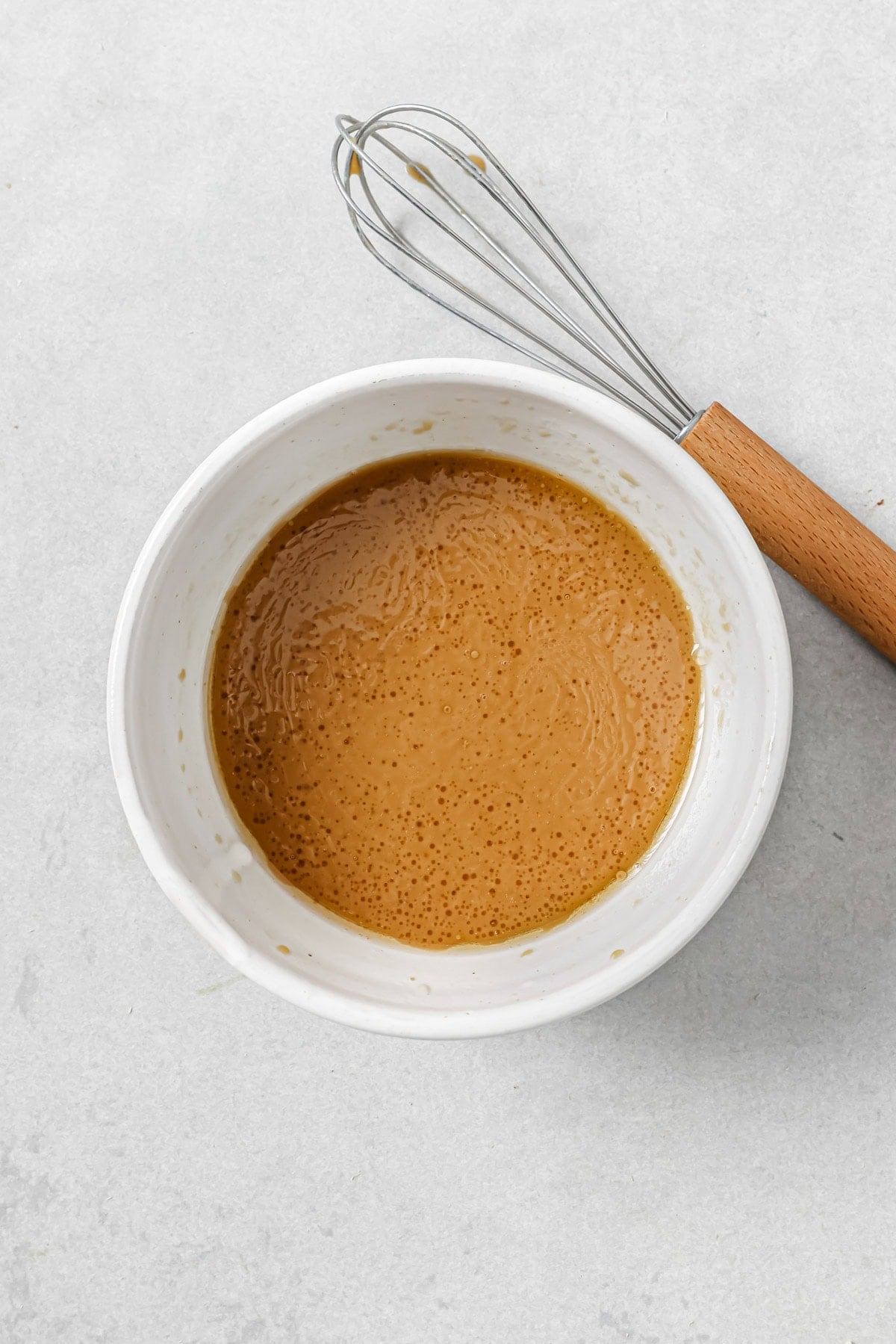 A white bowl filled with light brown, creamy sauce or batter sits on a light surface next to a metal whisk with a wooden handle.