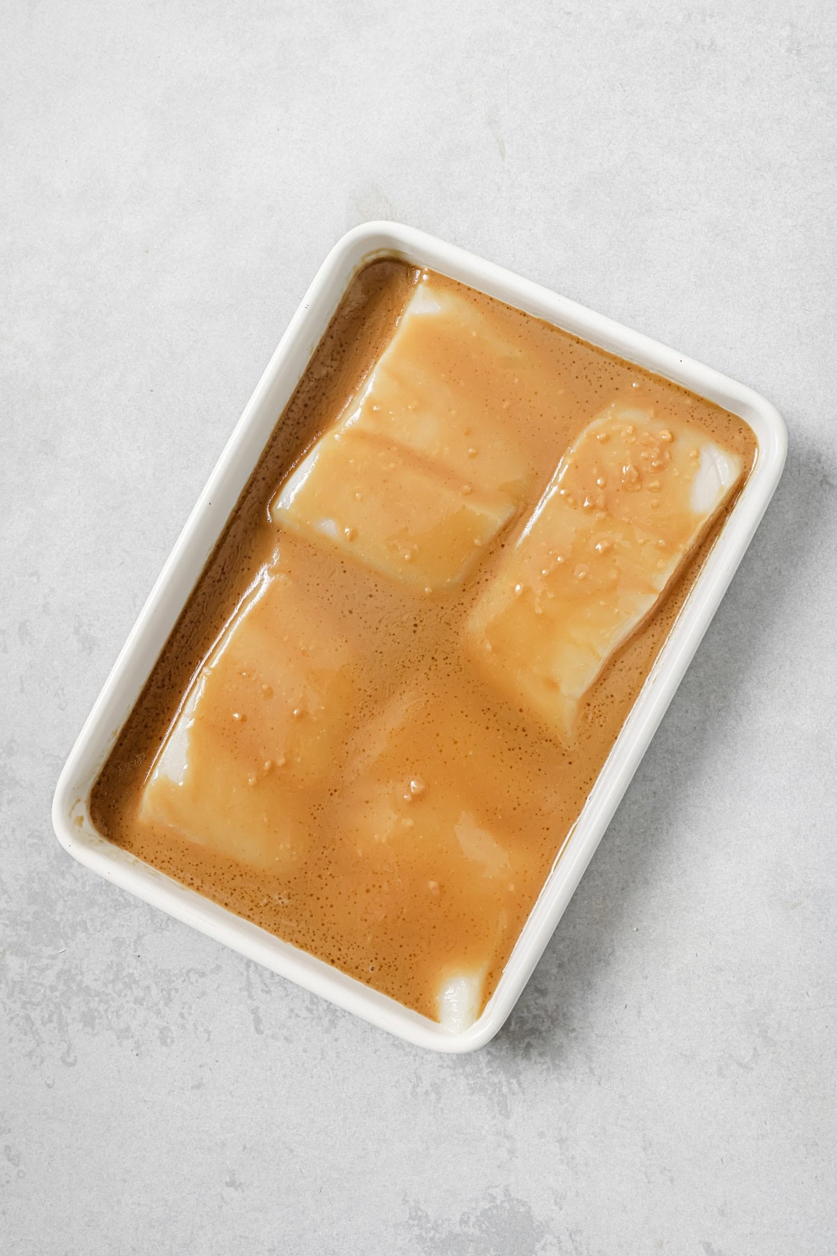 Four pieces of white fish sit in a rectangular white dish, covered with a light brown marinade on a light gray surface.