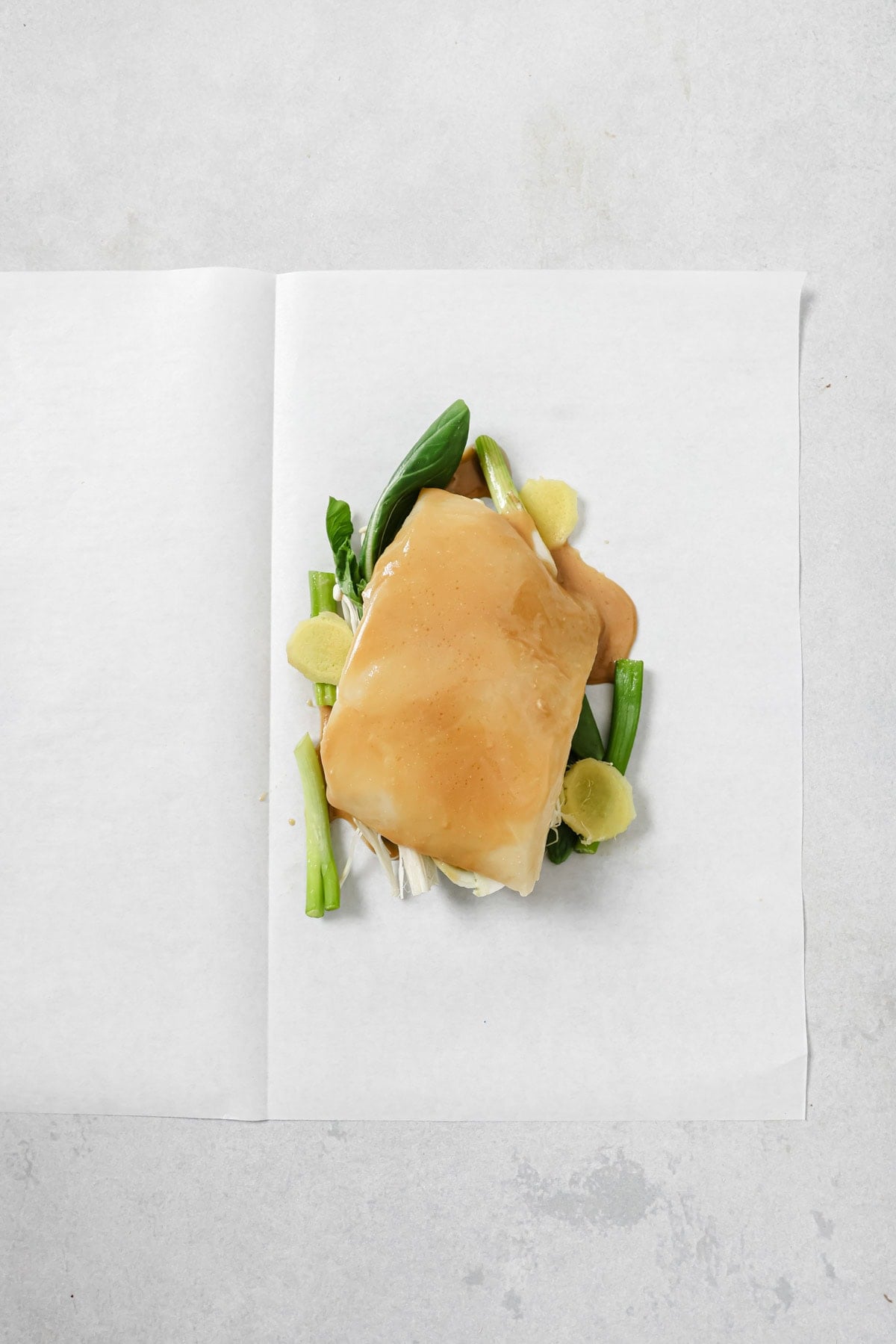 A parchment sheet holds a raw fish fillet topped with a light sauce, resting on green onions, spinach leaves, and ginger slices, ready for cooking.