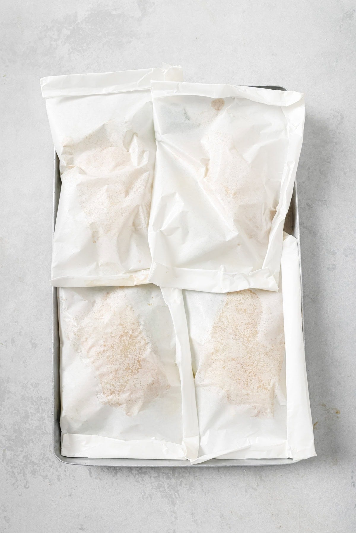 A baking tray holding four parchment paper packets, each containing food and sealed shut, placed on a light gray surface.