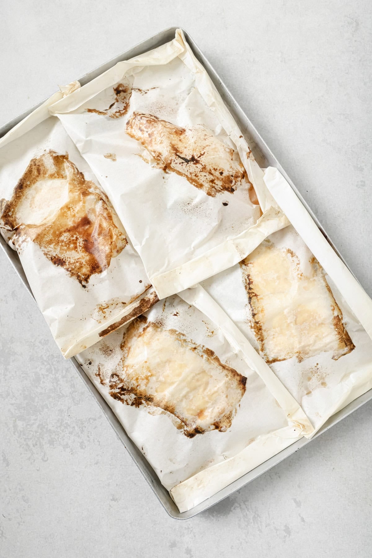 Four pieces of fish are baked in parchment paper packets on a metal baking tray, with some browning and juices visible on the parchment. The tray is placed on a light gray surface.