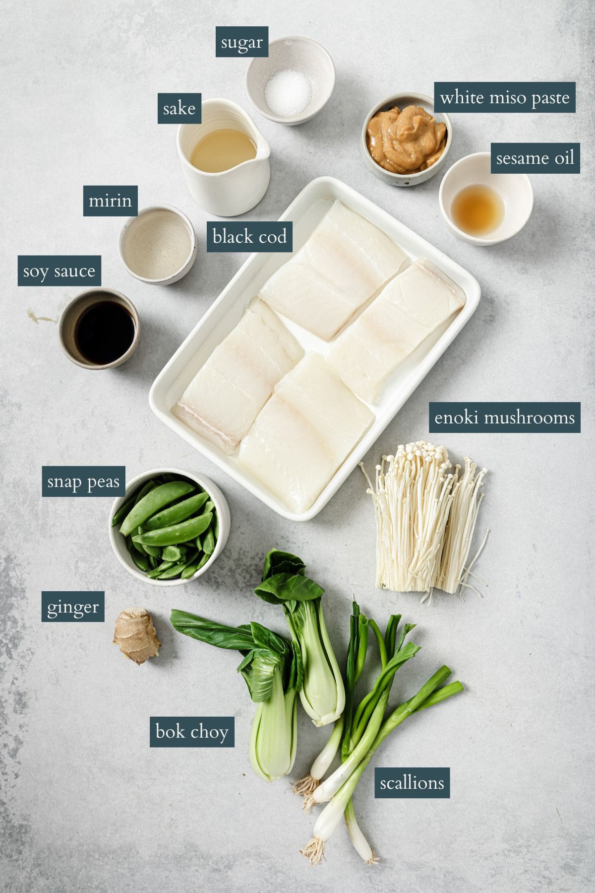 A flat lay of labeled ingredients for a recipe, including black cod fillets, white miso paste, sesame oil, enoki mushrooms, snap peas, soy sauce, sake, sugar, mirin, ginger, bok choy, and scallions on a light surface.