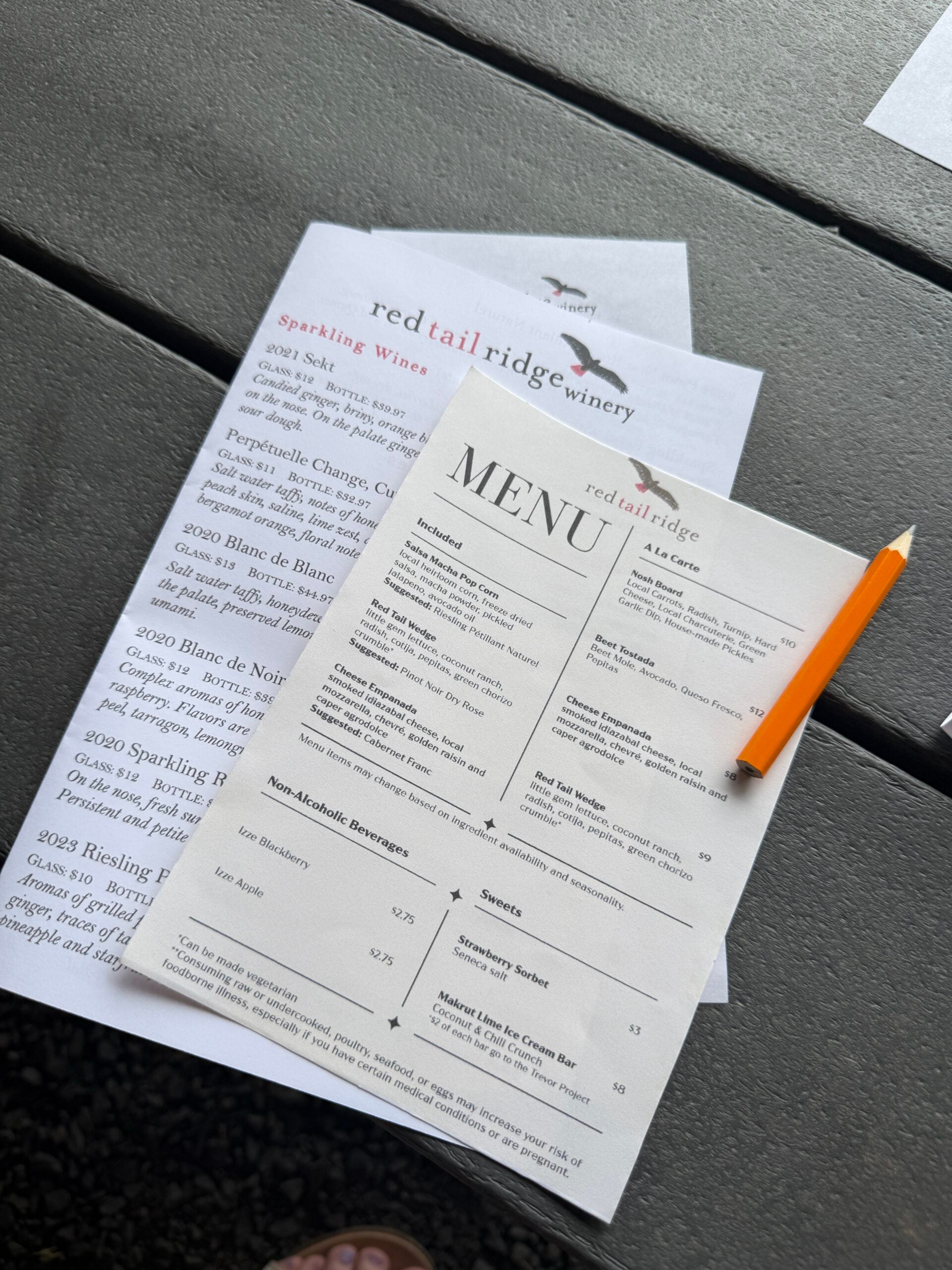 A pencil rests on top of two Red Tail Ridge Winery menus placed on a gray table, one featuring wine selections and the other listing food and non-alcoholic beverage options.