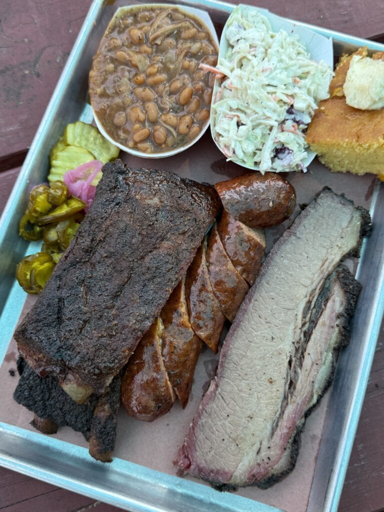 A tray with barbecue ribs, sausage, brisket, baked beans, coleslaw, cornbread, and pickles on parchment paper, served on a wooden table.