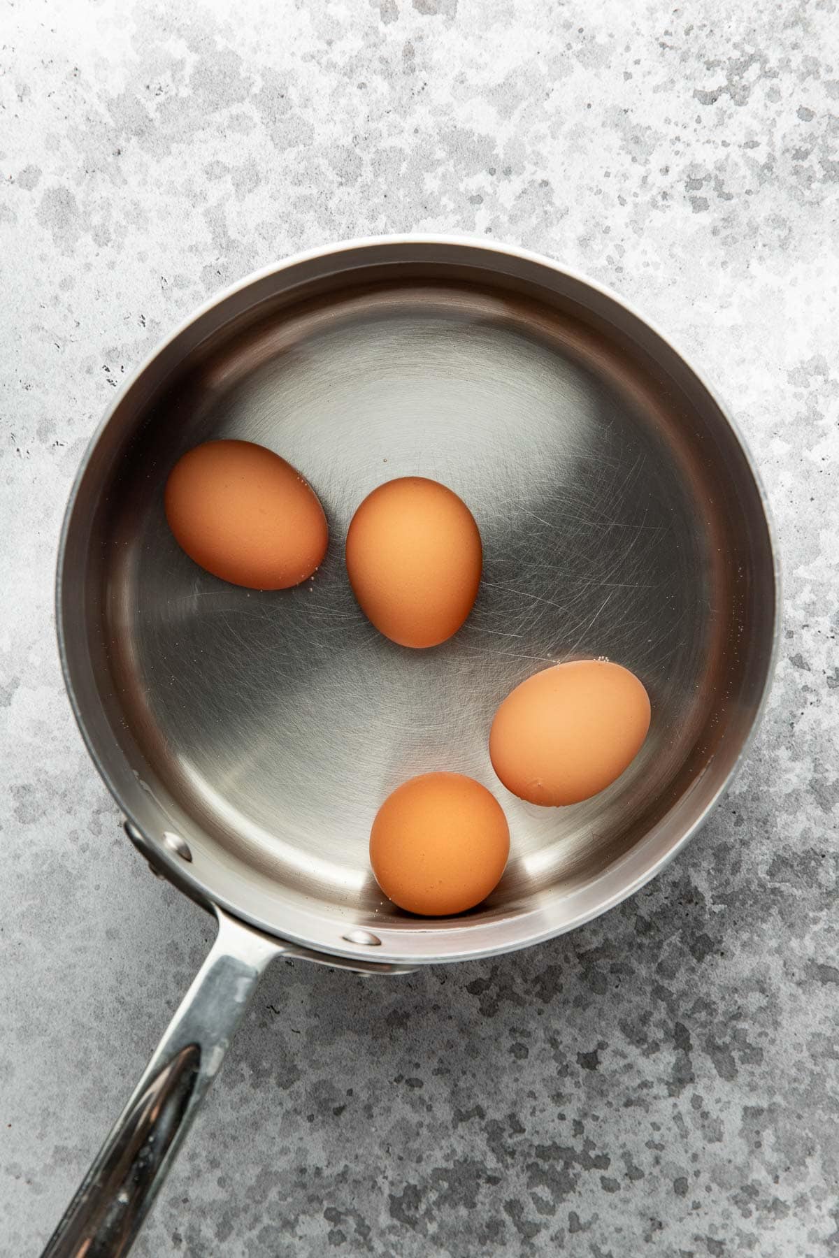 A stainless steel saucepan with four brown eggs inside, filled with water, sits on a gray textured surface.