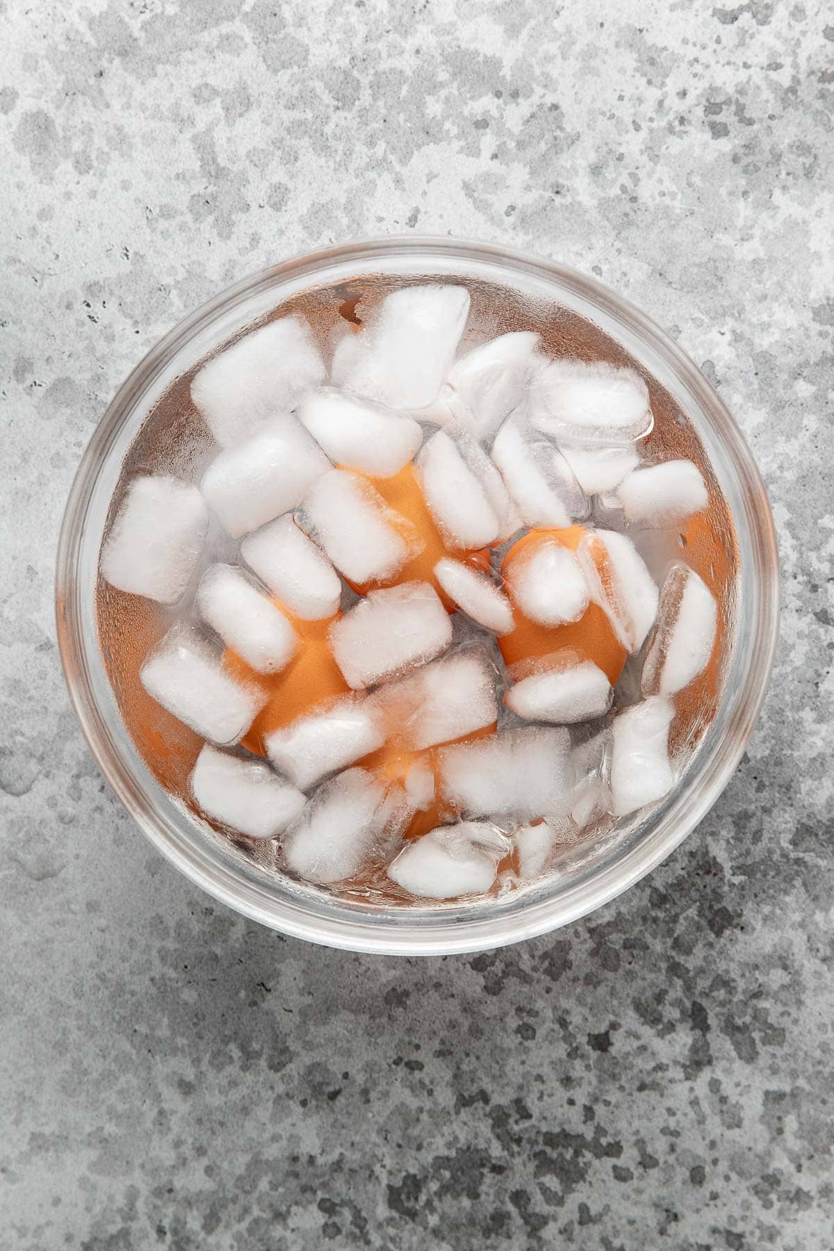 A glass bowl filled with ice cubes and brown eggs sits on a gray, textured surface.