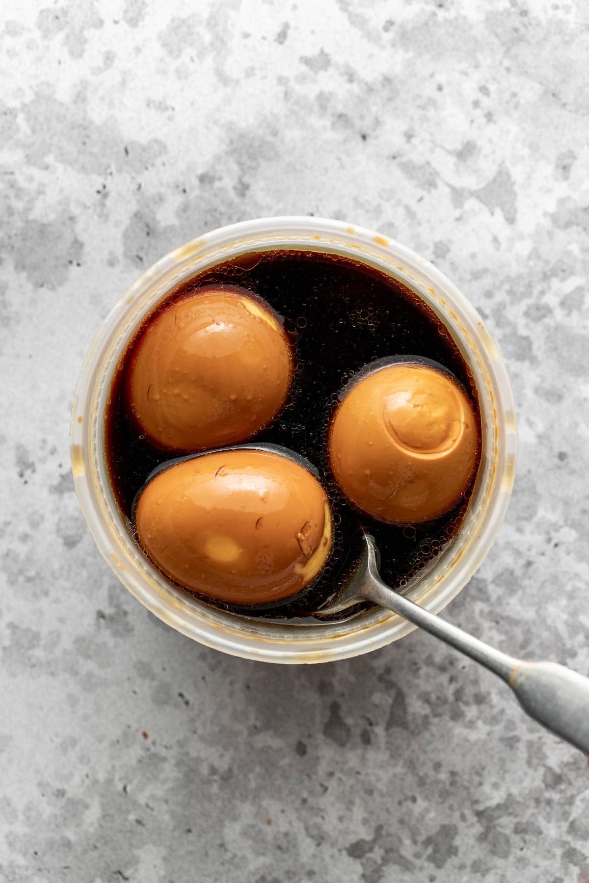 Three brown eggs soaking in dark soy sauce marinade in a round container, with a spoon resting inside. The container sits on a gray, textured surface.