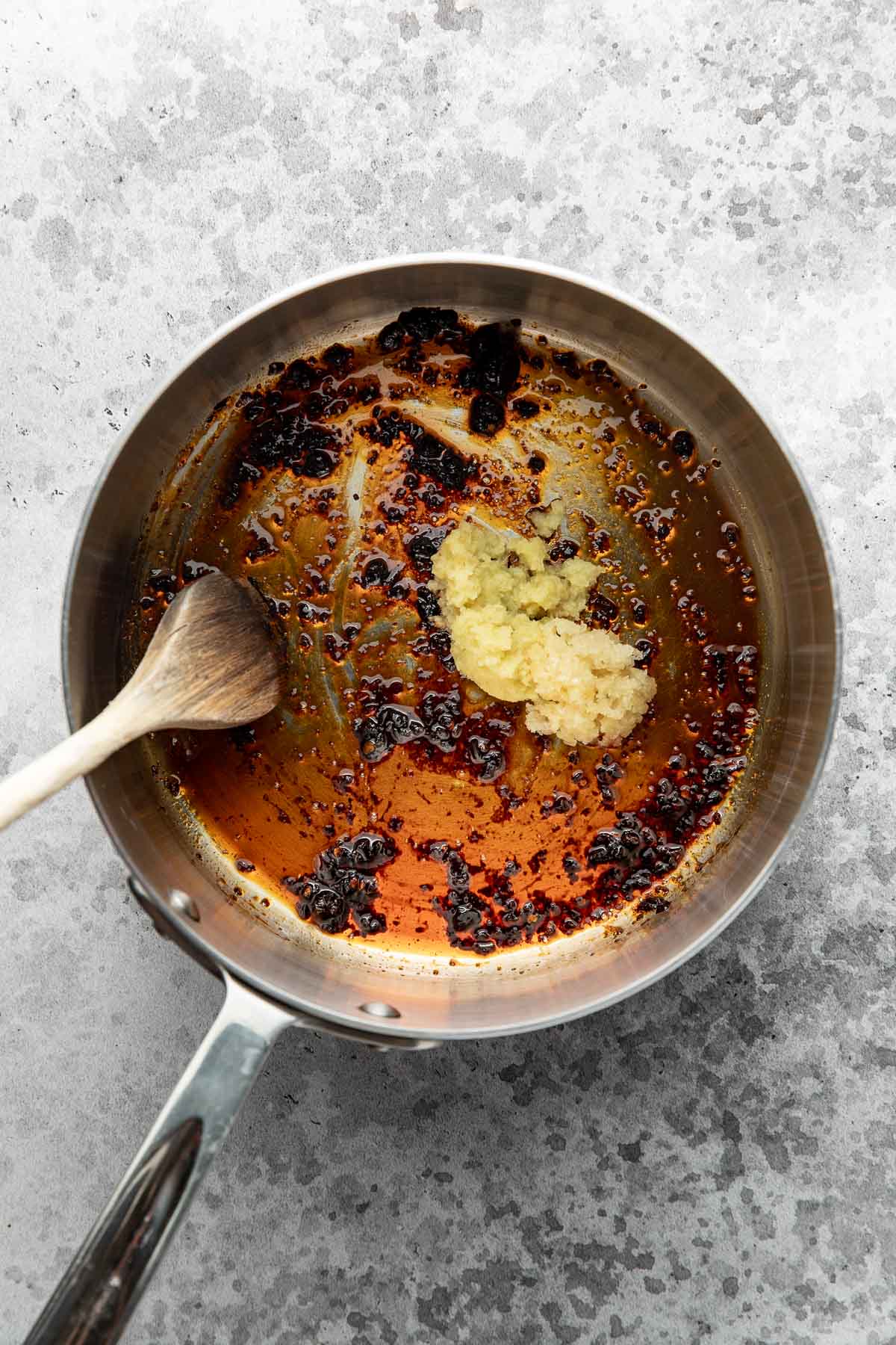 A saucepan with oil, dark chili flakes, and minced garlic being stirred with a wooden spoon on a gray textured surface.