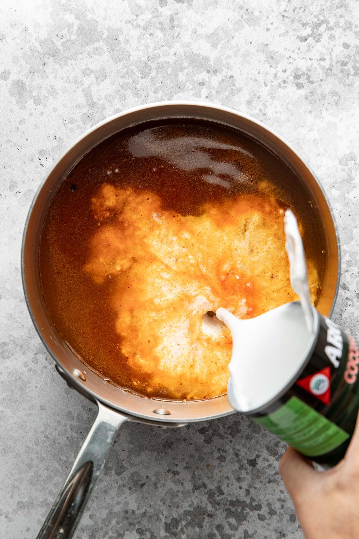 A hand pours coconut milk from a can into a saucepan filled with orange-colored broth, creating a swirling effect as the liquid mixes.