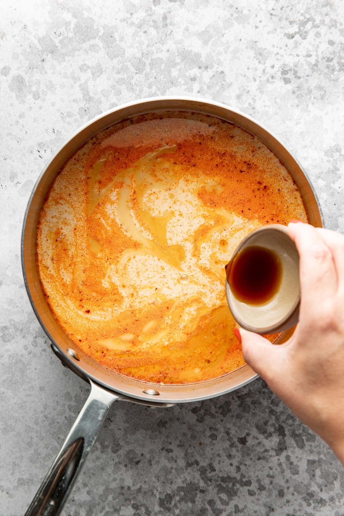 A hand pours dark liquid from a small cup into a saucepan filled with a swirling orange and cream-colored soup on a gray surface.