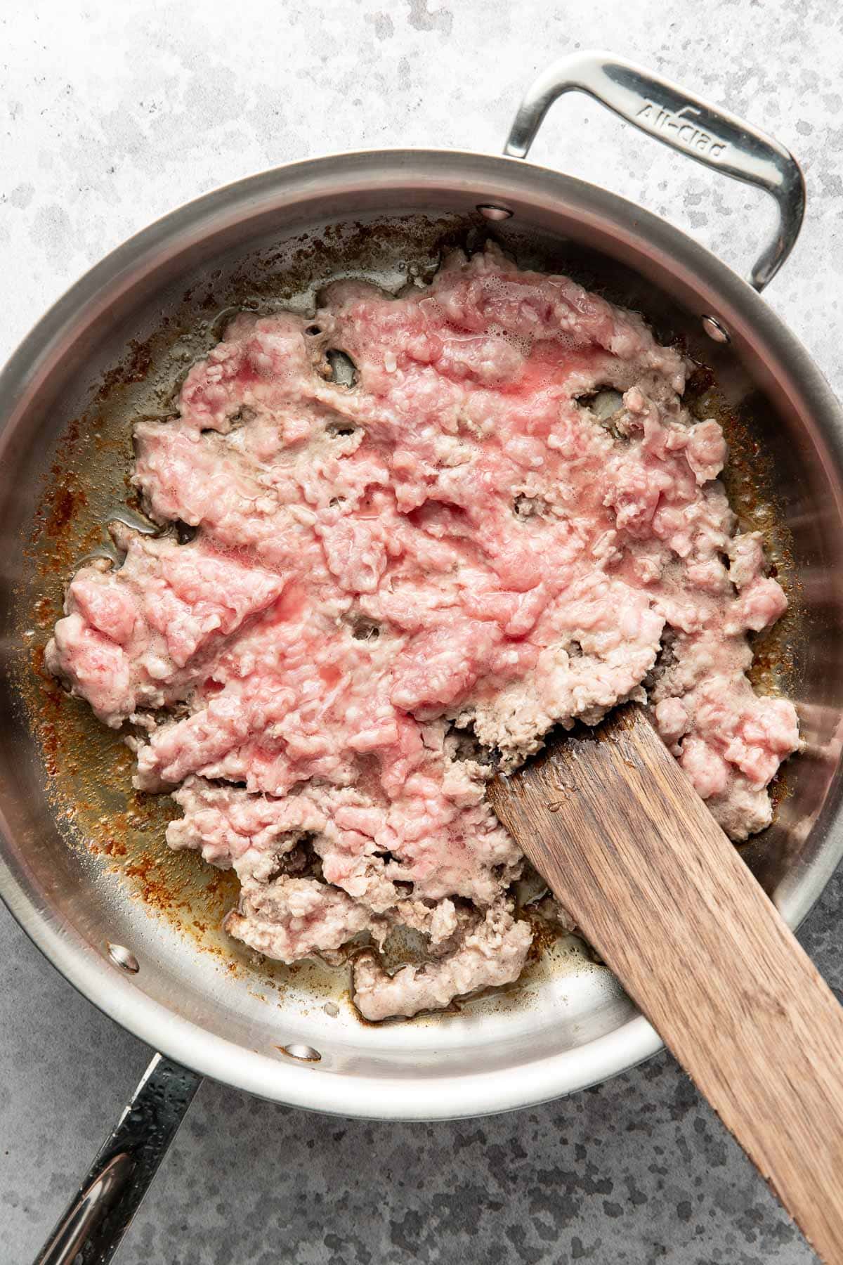 Ground meat cooking in a stainless steel skillet, partially browned, with a wooden spatula resting in the pan.