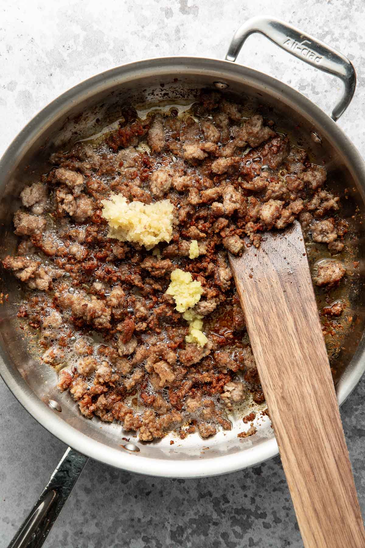 Ground sausage browning in a stainless steel pan with a wooden spatula and minced garlic added on top, ready to be mixed in.