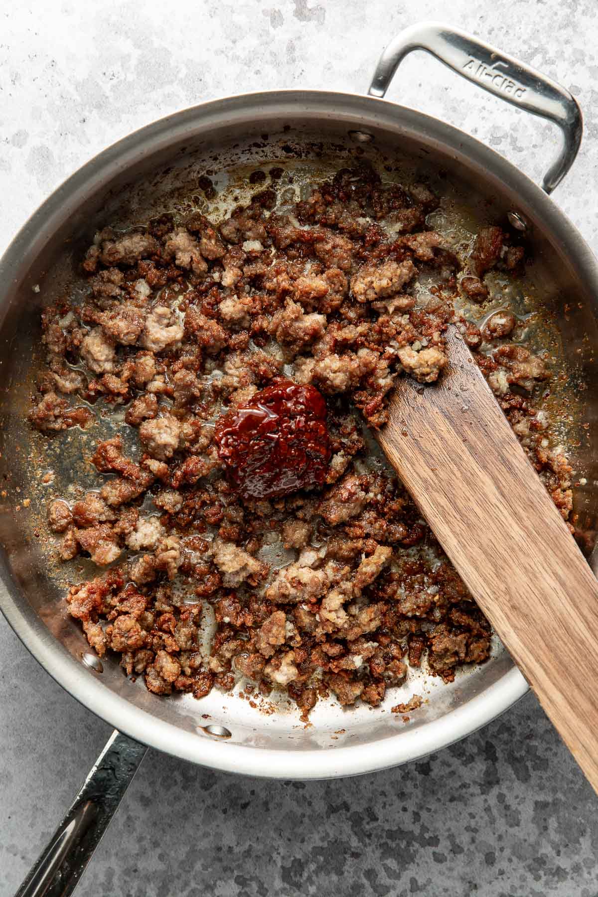 Cooked ground meat in a stainless steel skillet with a dollop of red sauce and a wooden spatula resting inside.