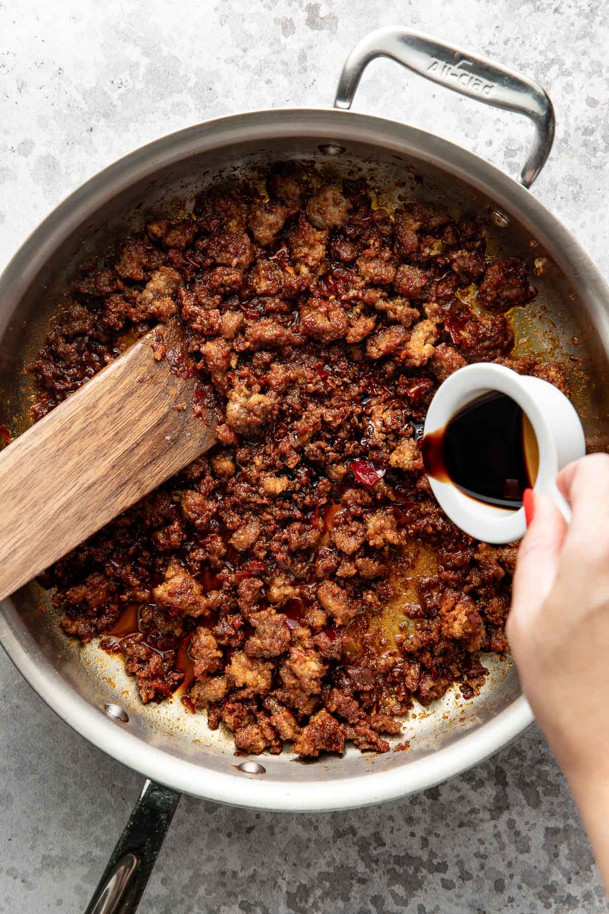 A hand pours a dark liquid from a small white cup into a pan of browned ground meat, next to a wooden spatula on a light countertop.
