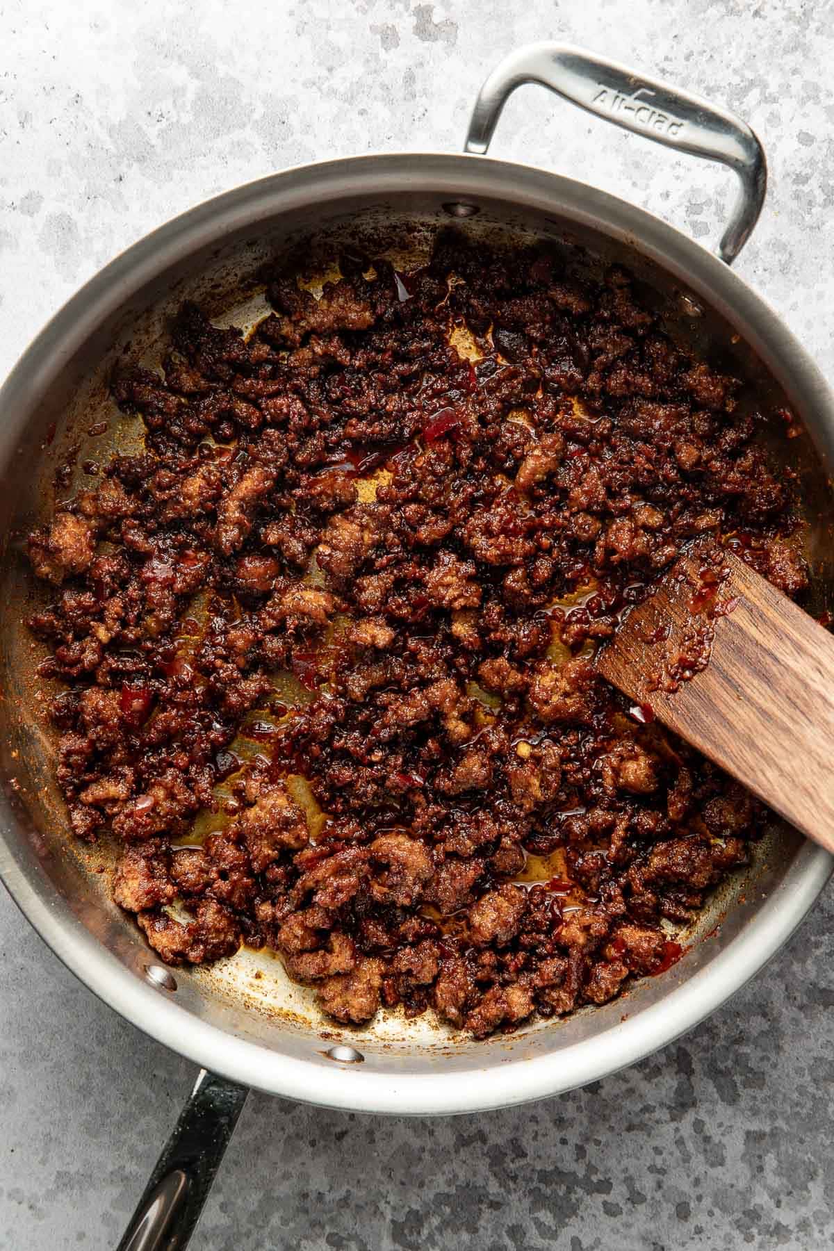 Cooked ground beef with seasoning in a stainless steel skillet, stirred with a wooden spatula. The meat appears browned and slightly caramelized, sitting in its juices on a grey surface background.