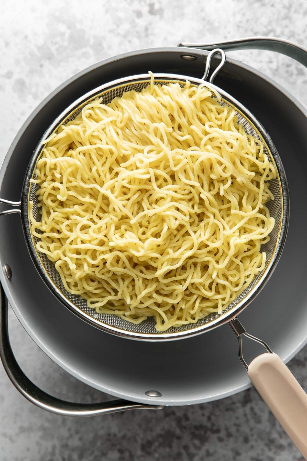 Cooked yellow noodles in a fine mesh strainer rest over a dark pot on a light countertop. The noodles appear drained and ready for use.
