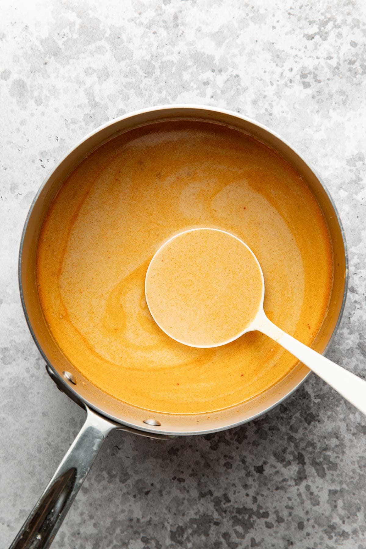 A saucepan filled with creamy, orange-colored sauce sits on a gray surface. A white ladle rests inside the pan, filled with the same smooth sauce.